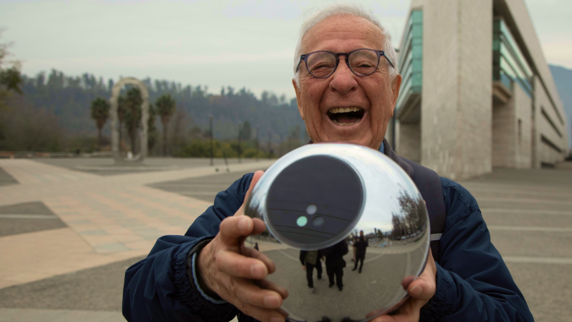 An elderly man joyfully holds the Orb, smiling in an outdoor setting with modern architecture in the background.