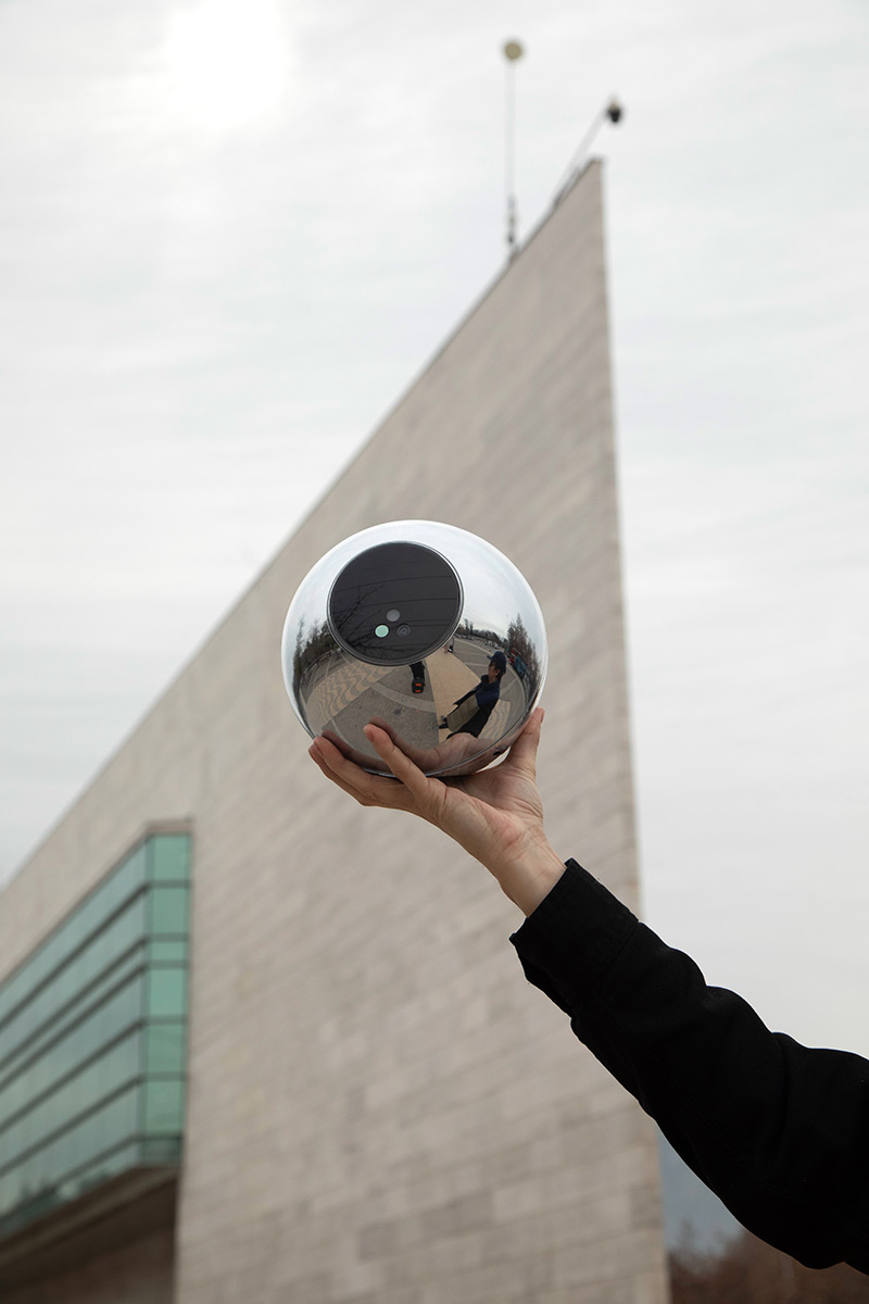 A person holds up the Orb against a modern building backdrop, reflecting their surroundings.