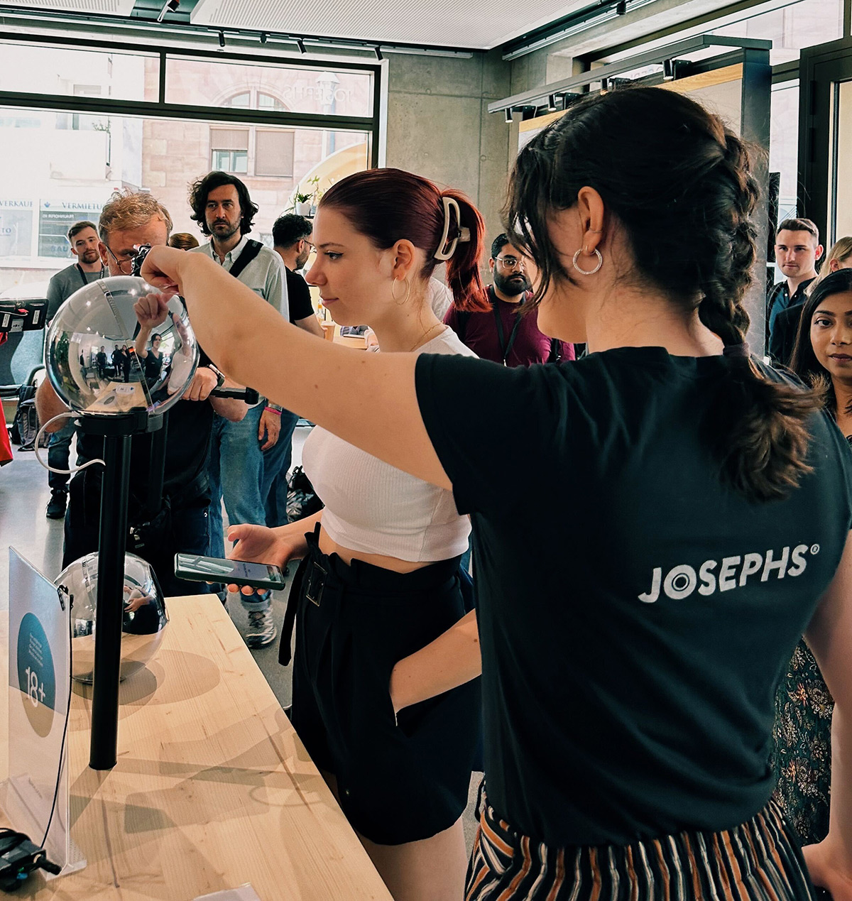 A person using the Orb for verification, while another assists in a lively environment with onlookers. People are engaged and interested.