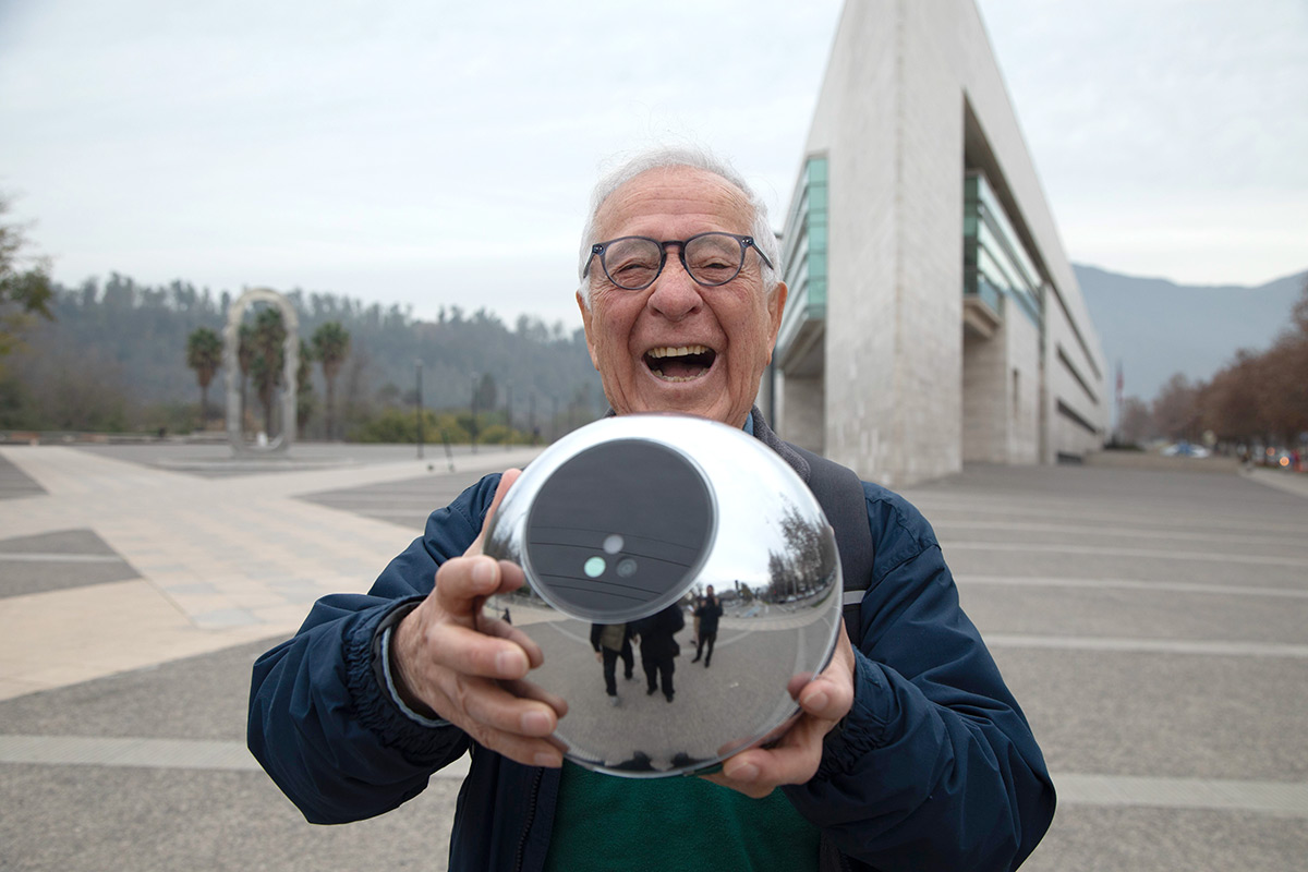 An elderly man joyfully holds an Orb in front of a modern building, with reflections of people and trees visible in the Orb.