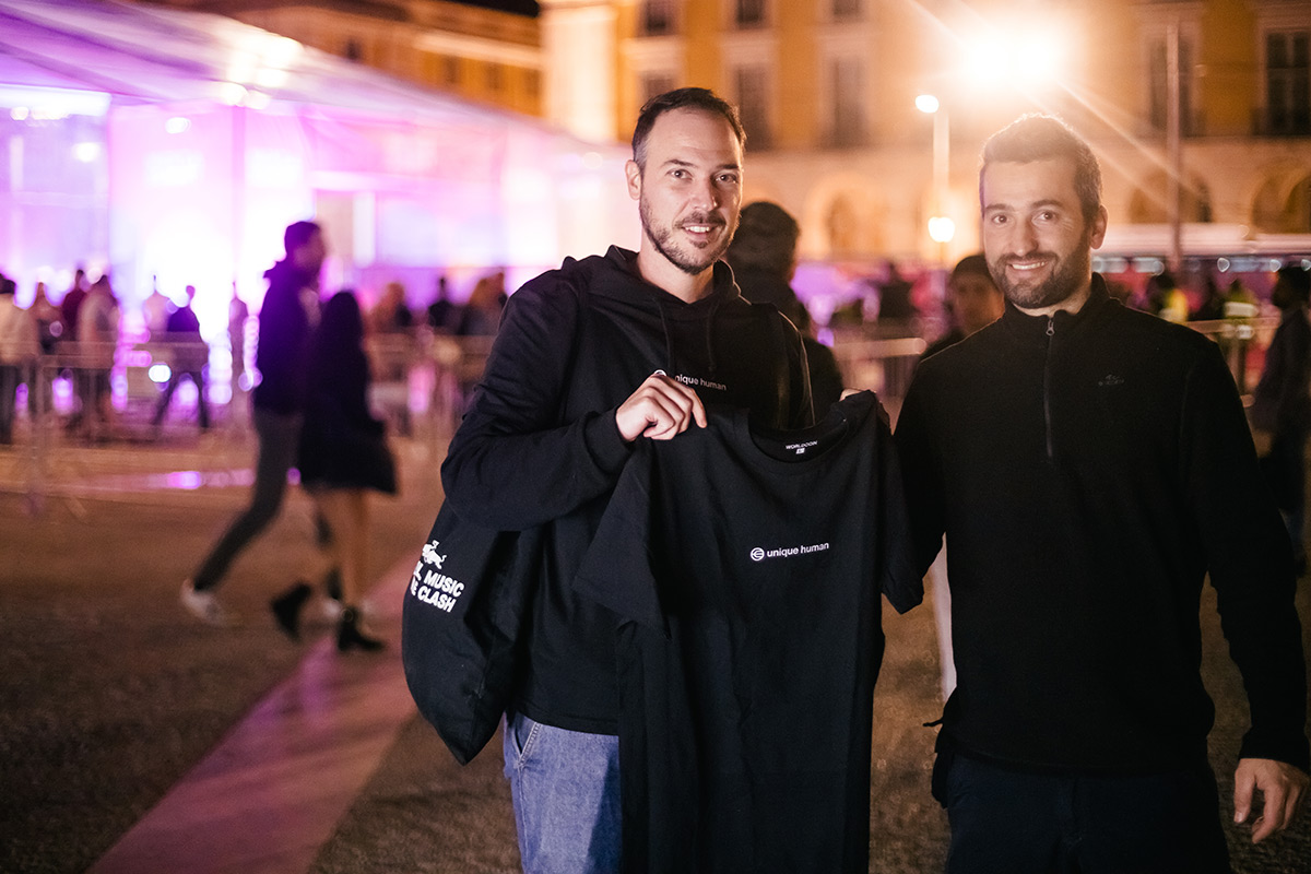 Two people smiling and holding a black t-shirt with a logo, standing in a lively outdoor setting with illuminated structures in the background.