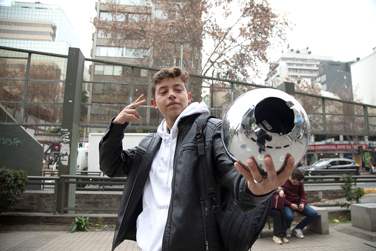 A young person poses confidently with an Orb in their hand, smiling and making a peace sign. In the background, two people sit relaxingly, surrounded by urban scenery.