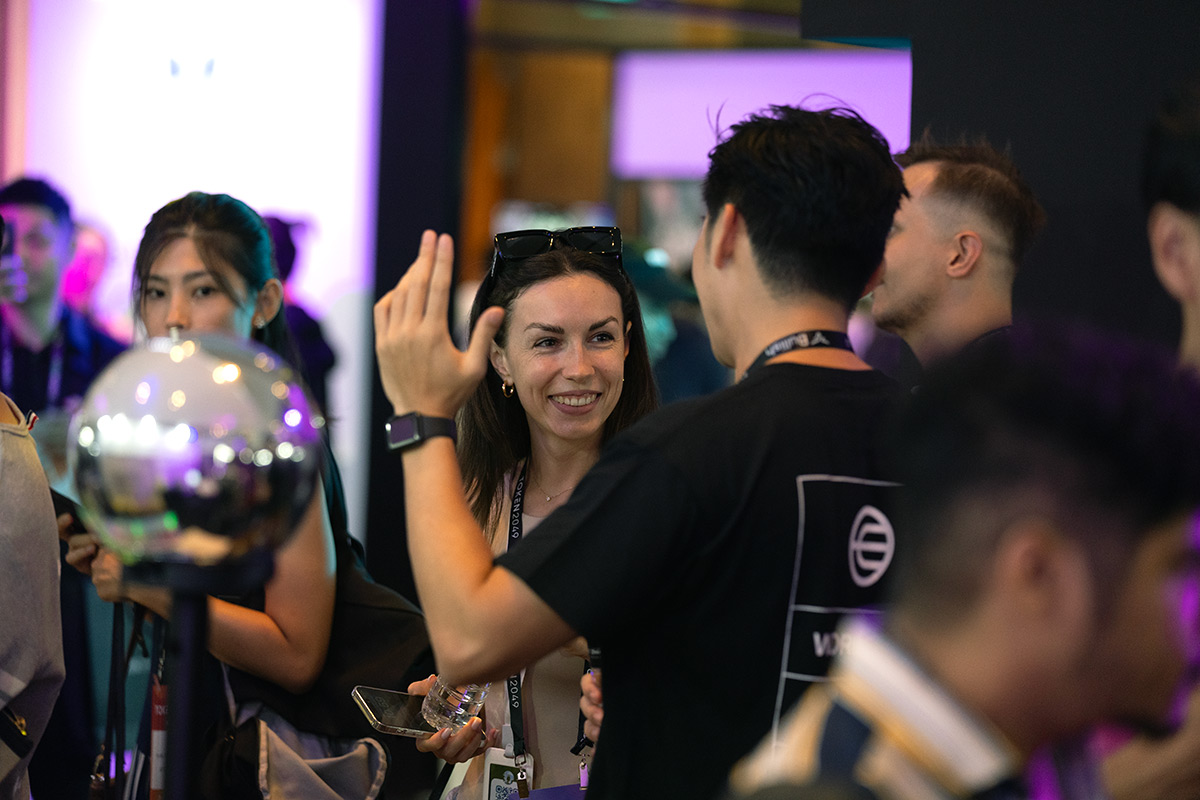 People engaging and interacting at an event, with one woman smiling and using her phone while a man gestures animatedly nearby. An Orb is visible in the foreground, reflecting the vibrant event atmosphere.