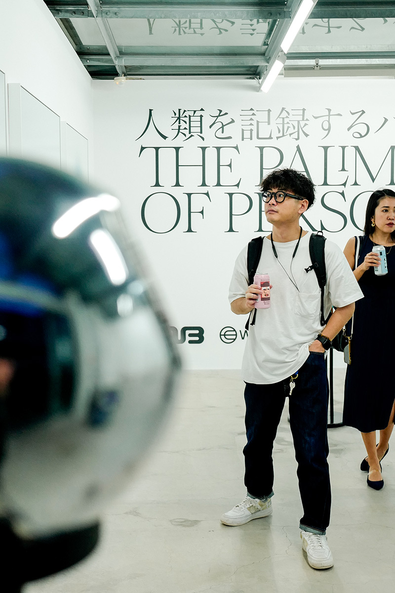 A person stands attentively with a drink, looking at a futuristic device in a gallery setting. Text in the background reads, 'THE PALM OF PERSON.' The environment feels modern and engaging.