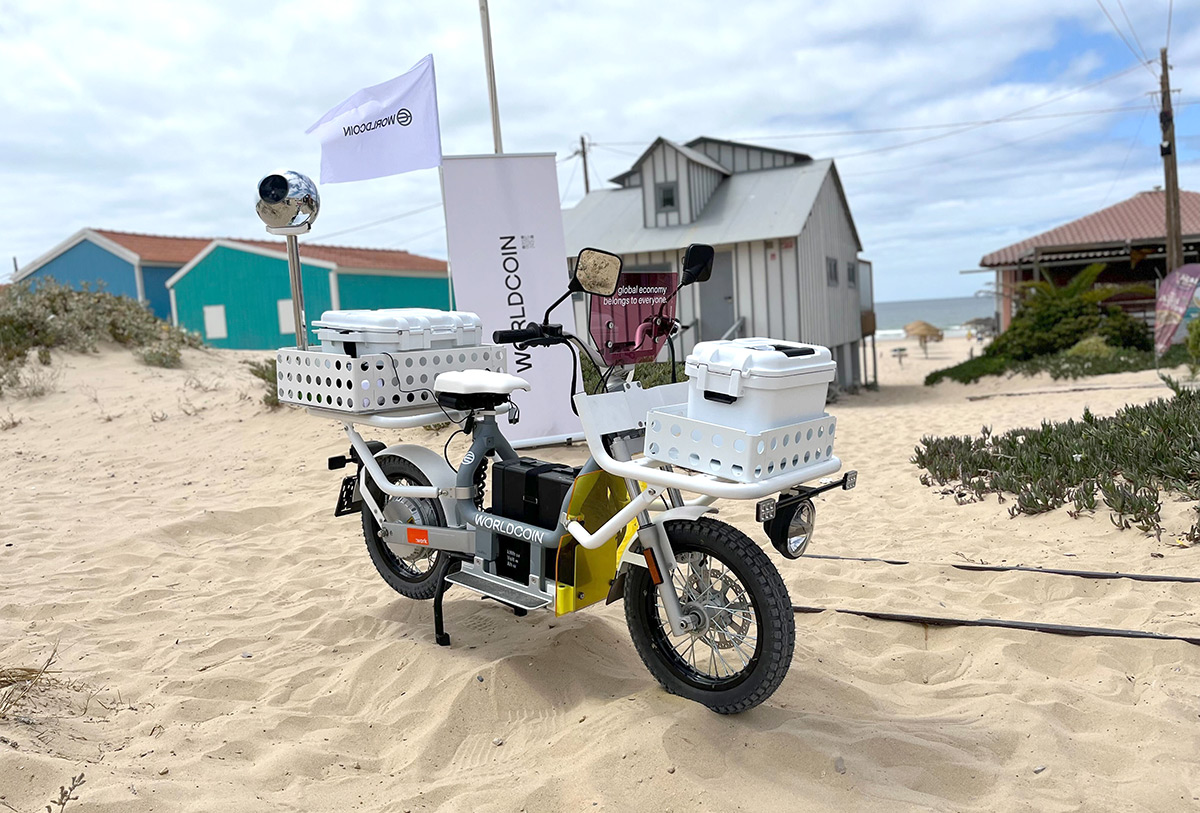 An electric bike with baskets is parked on sandy beach terrain, with colorful beach houses in the background. A Worldcoin banner stands beside the bike, and a silver Orb is mounted on the front.