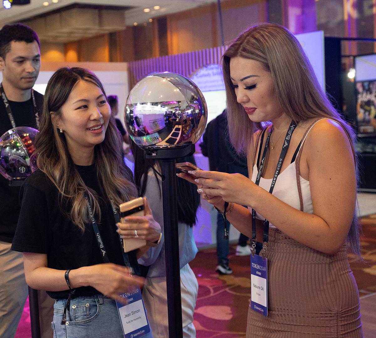 Two women interacting at an event, one using a smartphone while the other smiles near a shiny Orb on a stand.