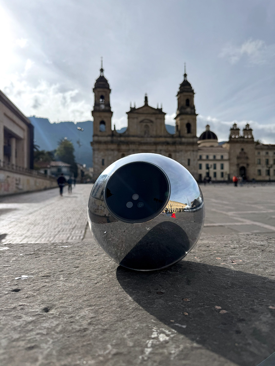 A silver Orb rests on a stone surface in a plaza, with historic buildings and mountains in the background under a partly cloudy sky.