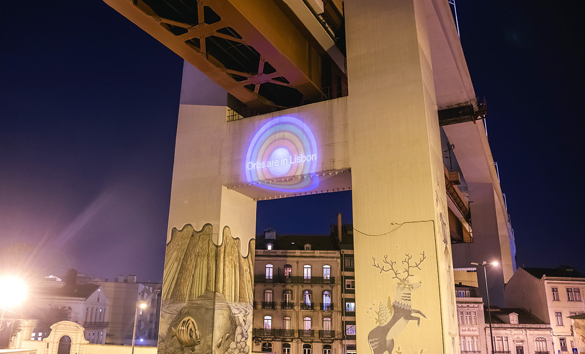A vibrant urban scene at night featuring a bridge with a colorful projection that says 'Orbs are in Lisbon' on a concrete pillar. The surrounding buildings are illuminated, enhancing the lively atmosphere.