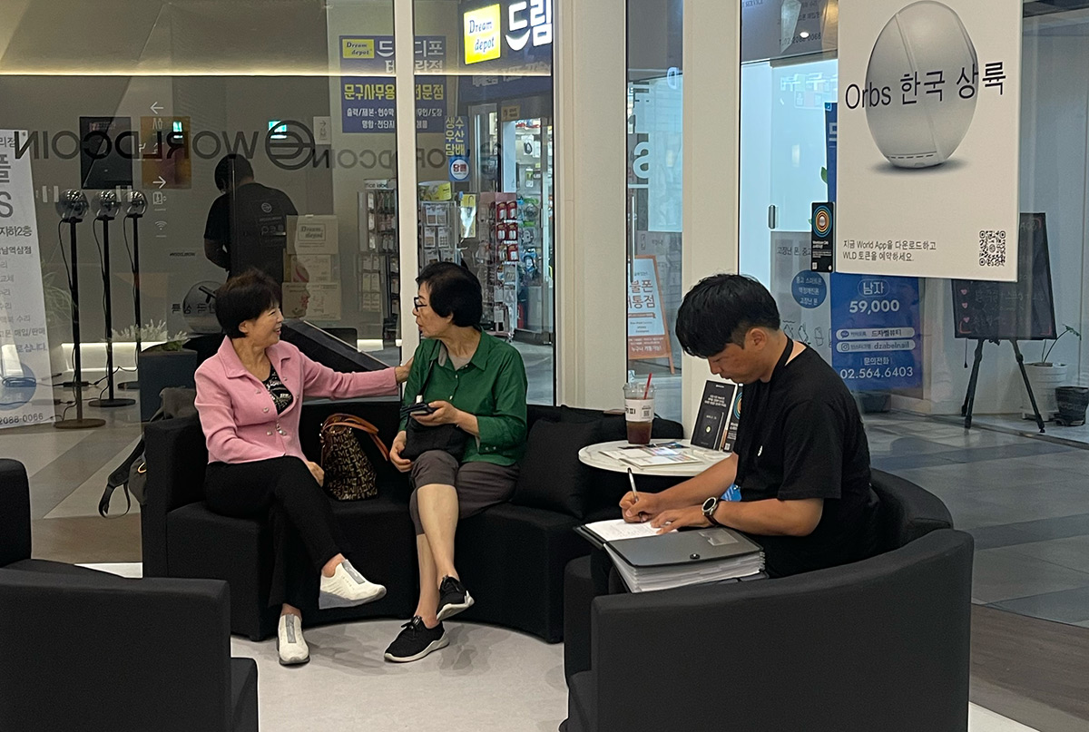 Two women engaged in conversation while sitting on a couch in a modern space, with a man nearby taking notes. An Orb device is visible in the background, promoting biometric verification.