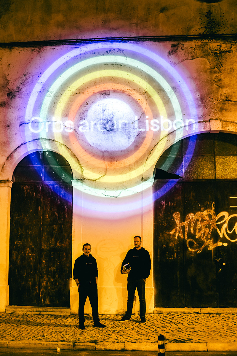 Two individuals stand in front of a colorful projection displaying concentric circles and text on a wall. The scene is illuminated at night, creating an engaging atmosphere.