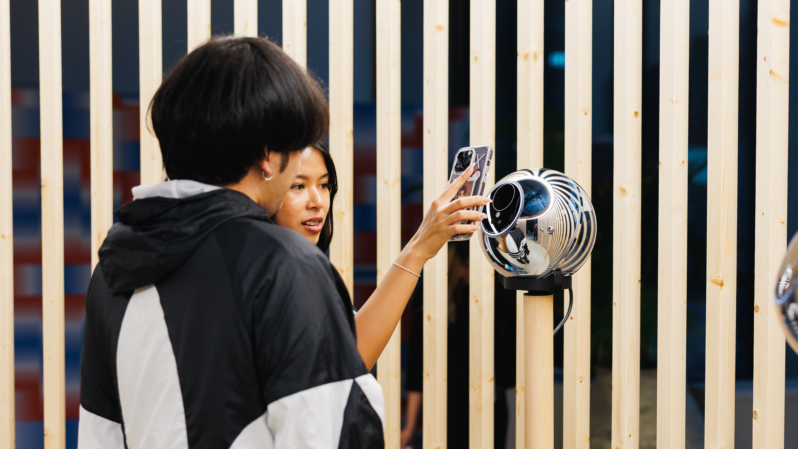 A person using a smartphone to verify with the Orb, while another person looks on, in a setting with wooden slats.