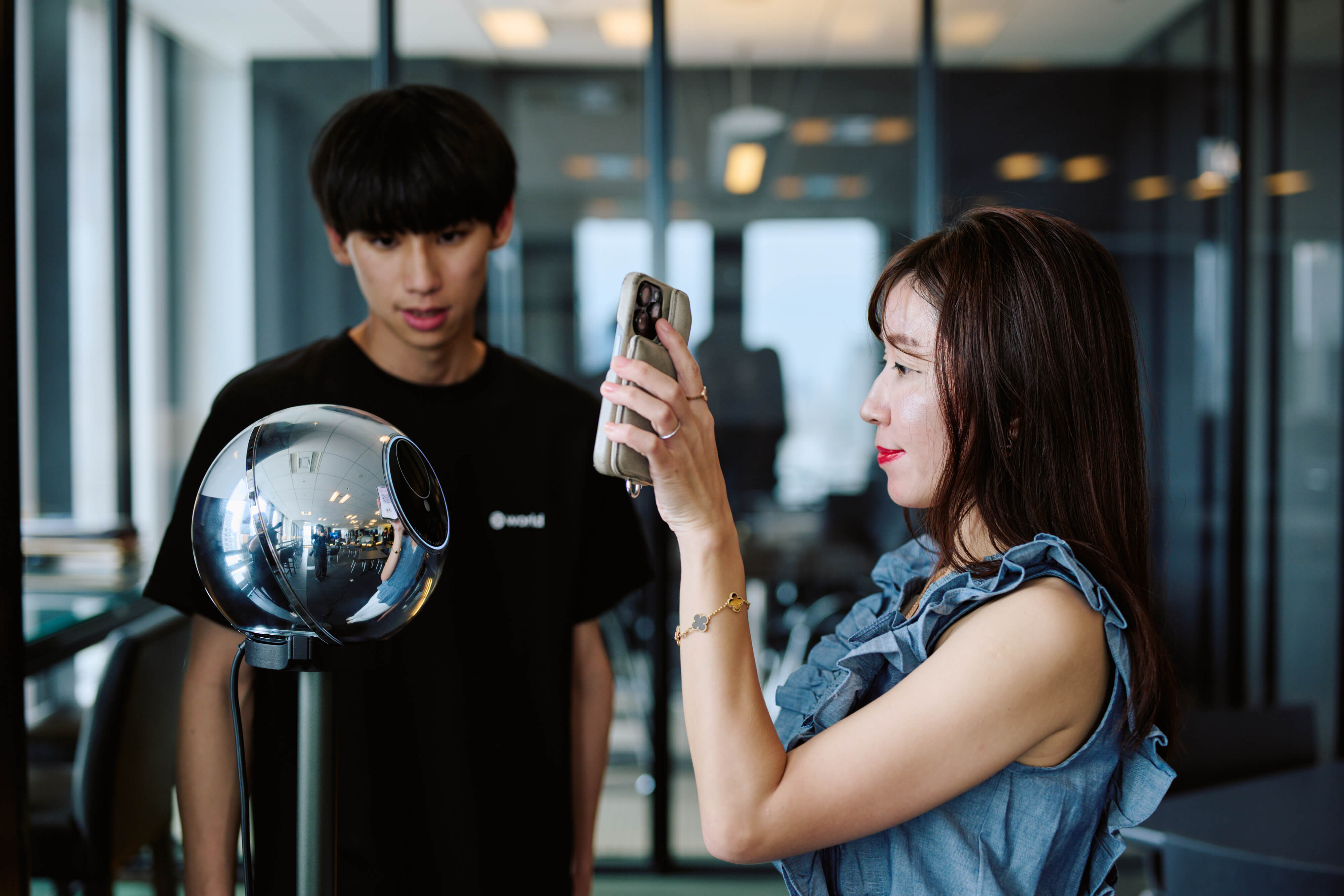 A woman using her phone to take a picture of her verification process with an Orb, while a man looks on with interest.