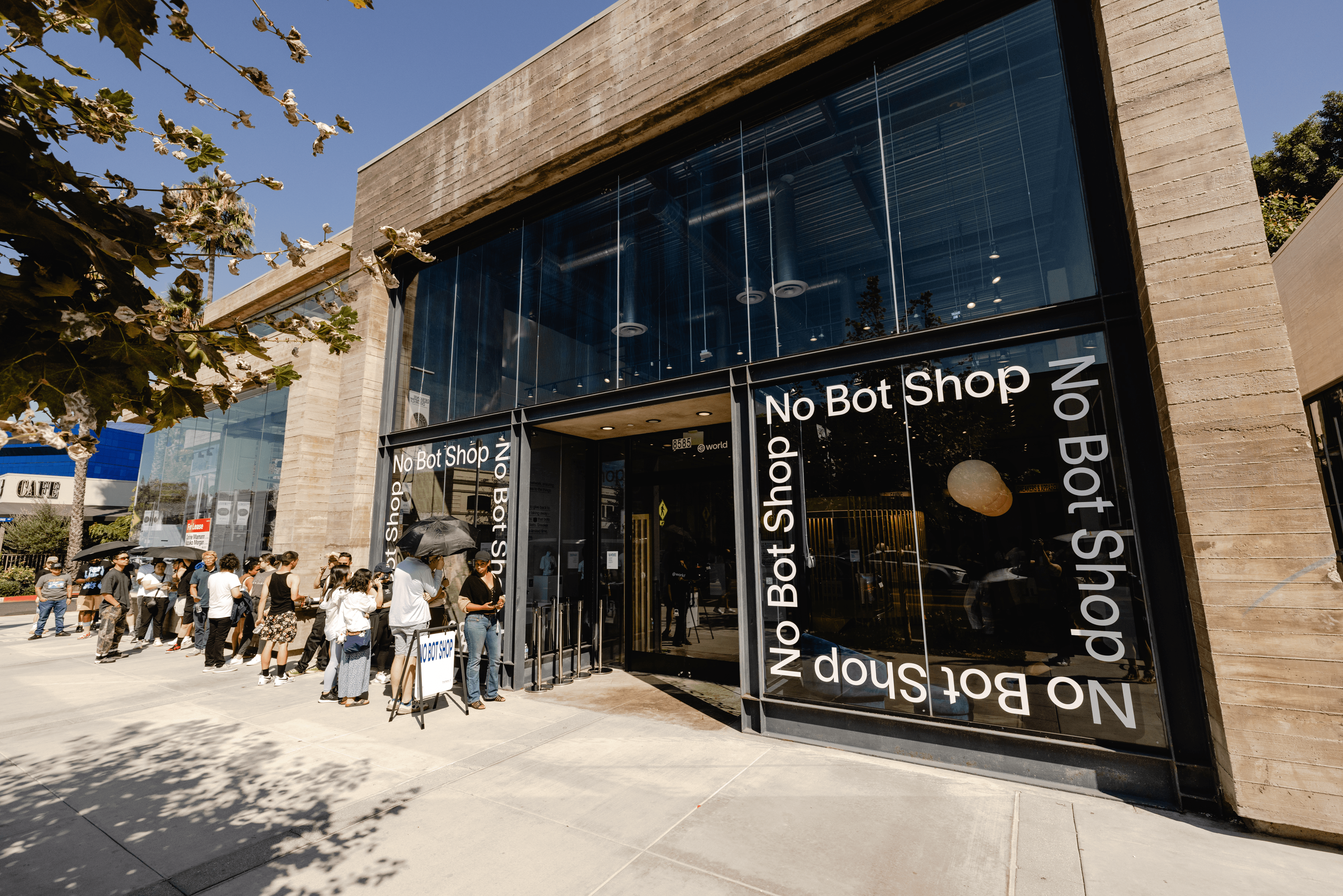 Exterior of the No Bot Shop with a line of people waiting to enter, featuring large windows with the text 'No Bot Shop.'