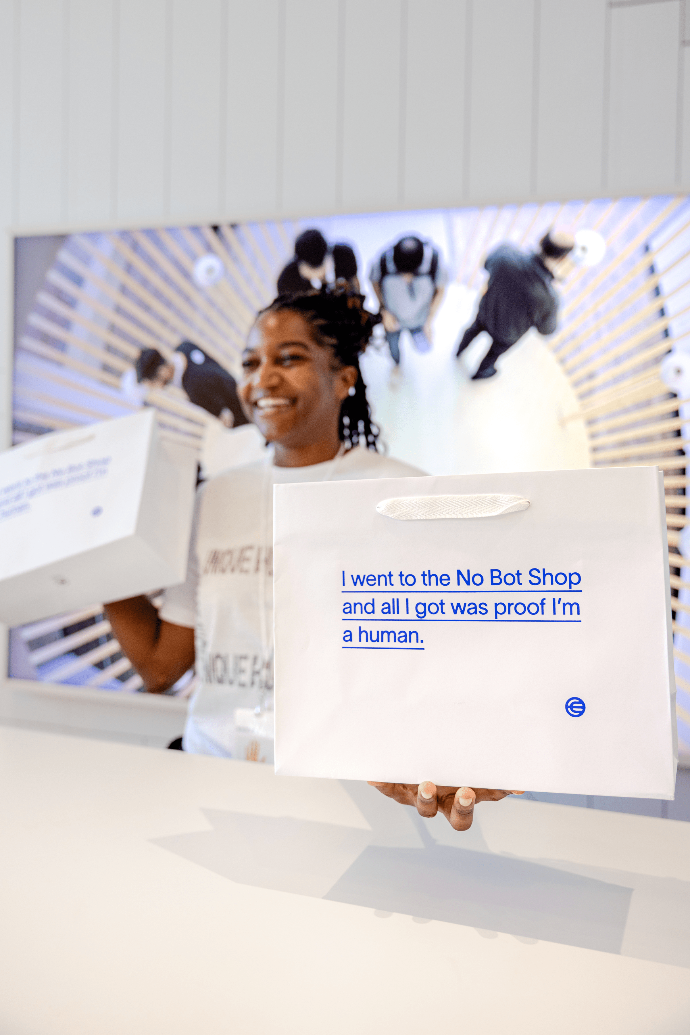 A smiling person holding a white bag with blue text that reads, 'I went to the No Bot Shop and all I got was proof I'm a human.' The background features a circular design with people visible in the distance.