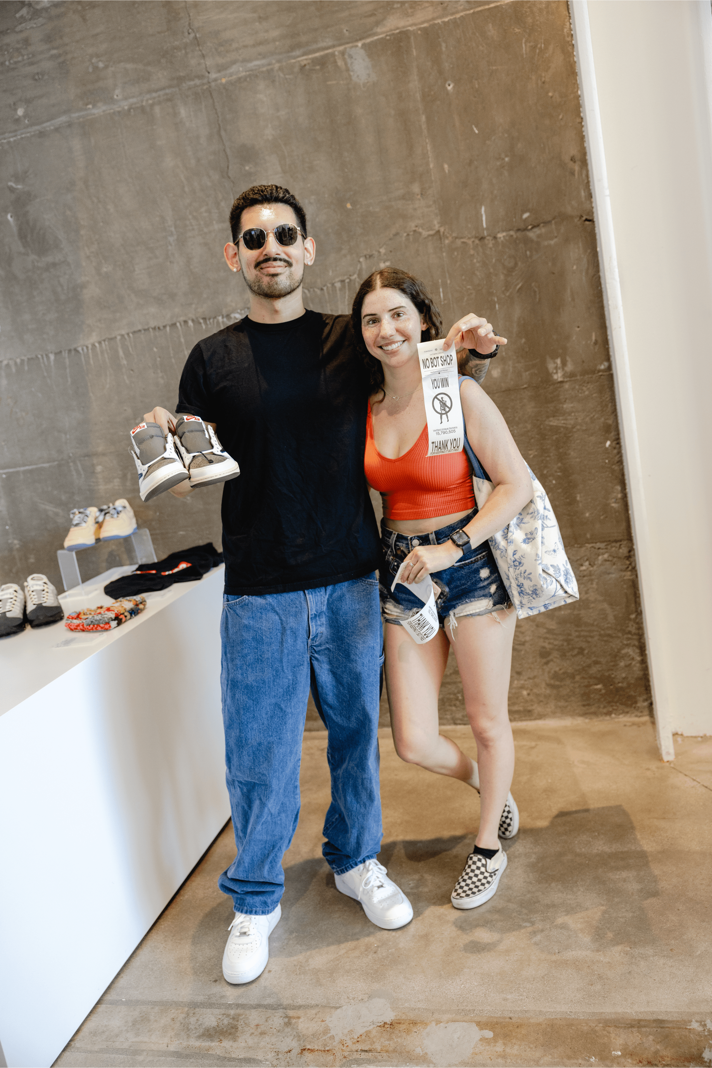 A man and woman pose for a photo, the man holding a pair of shoes while smiling, and the woman stands beside him also smiling. They are surrounded by sneakers on display in a stylish indoor setting.