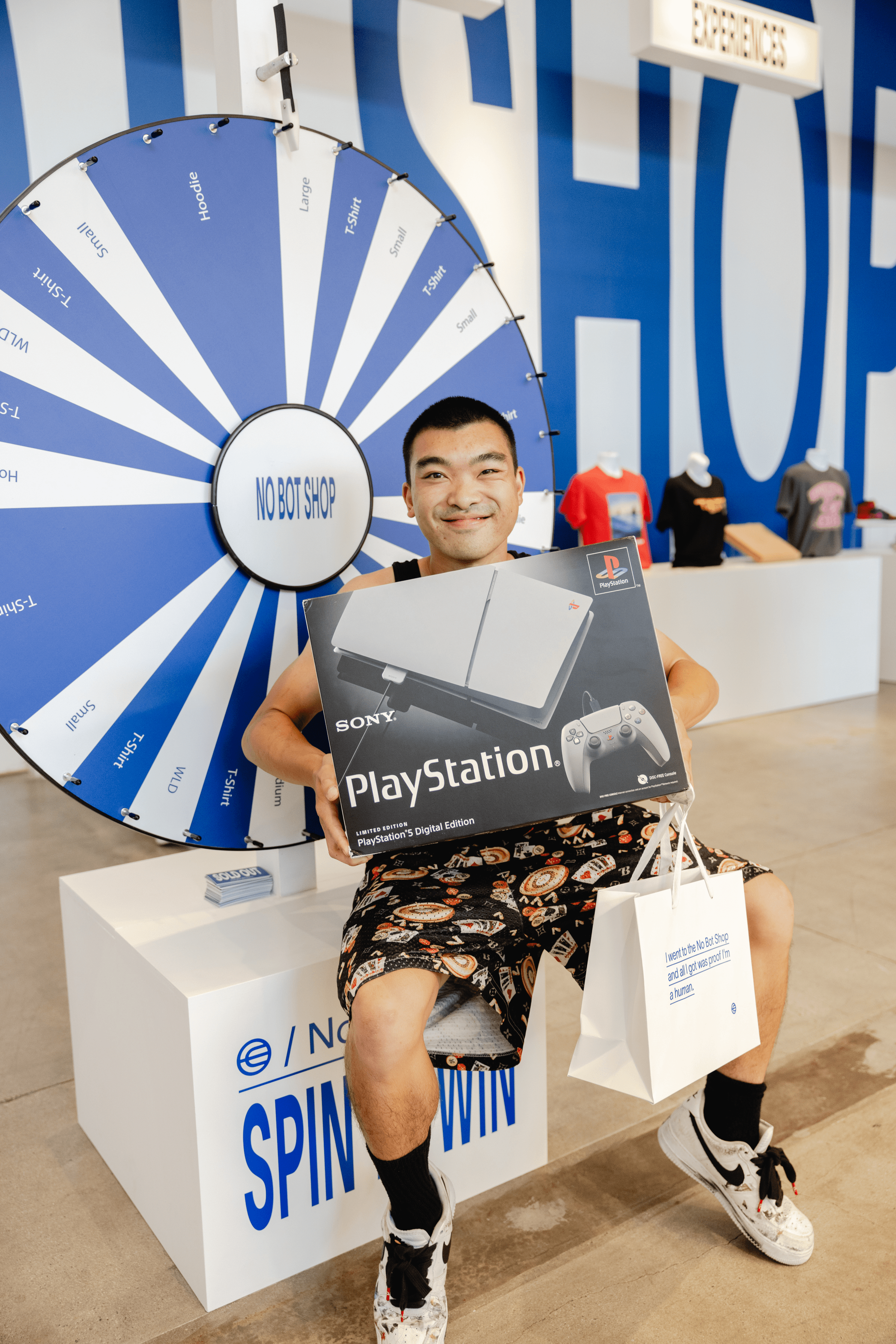 A smiling person holds a boxed PlayStation console while sitting next to a spinning prize wheel at a promotional event.
