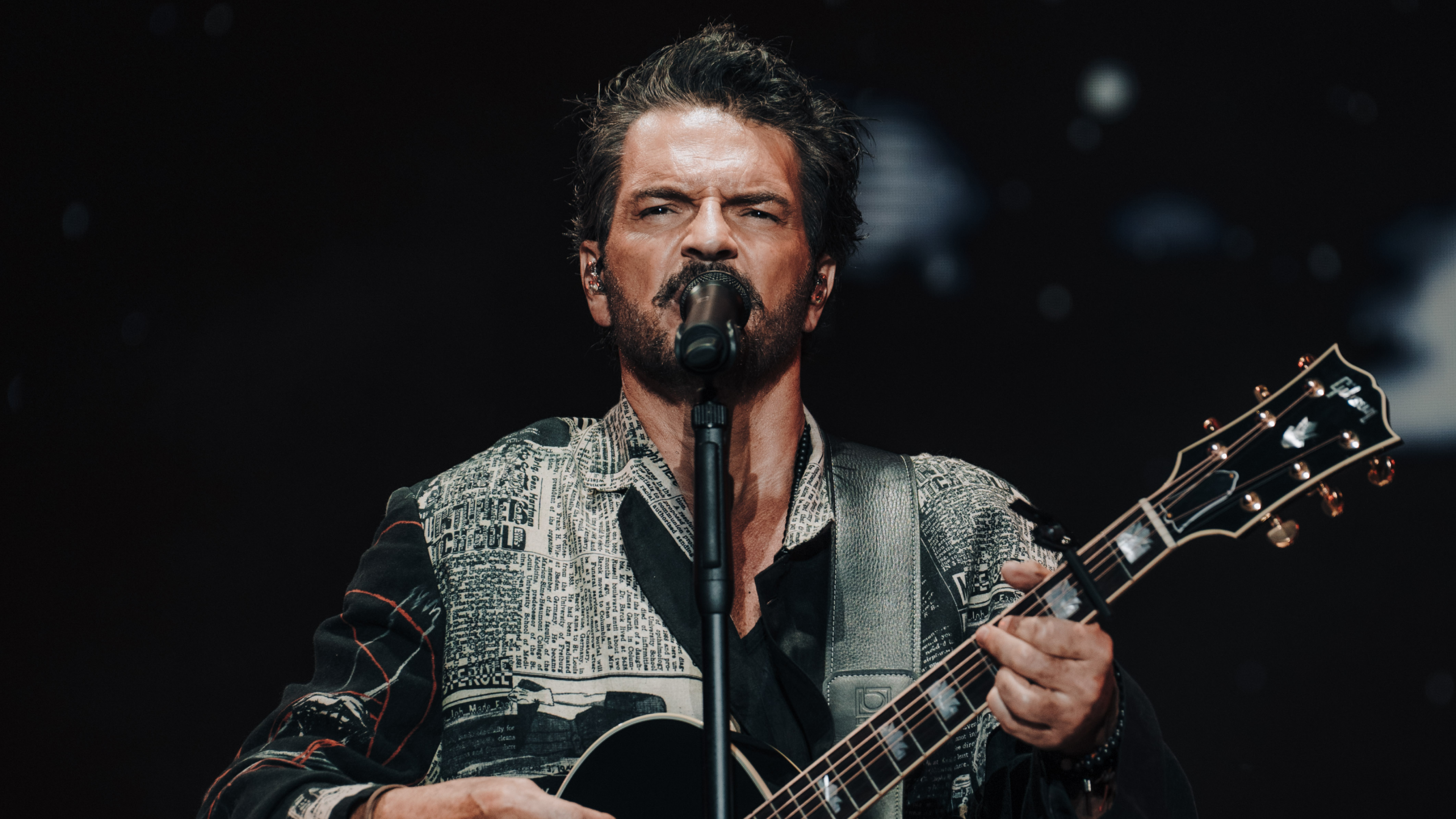 A performer singing into a microphone while playing an acoustic guitar, dressed in a stylish patterned outfit, engaging the audience.