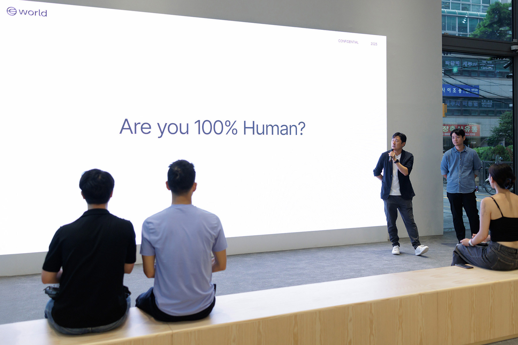 Un orador se presenta en un evento con una pantalla grande con la pregunta “¿Eres 100 % humano?” mientras una audiencia escucha atentamente.