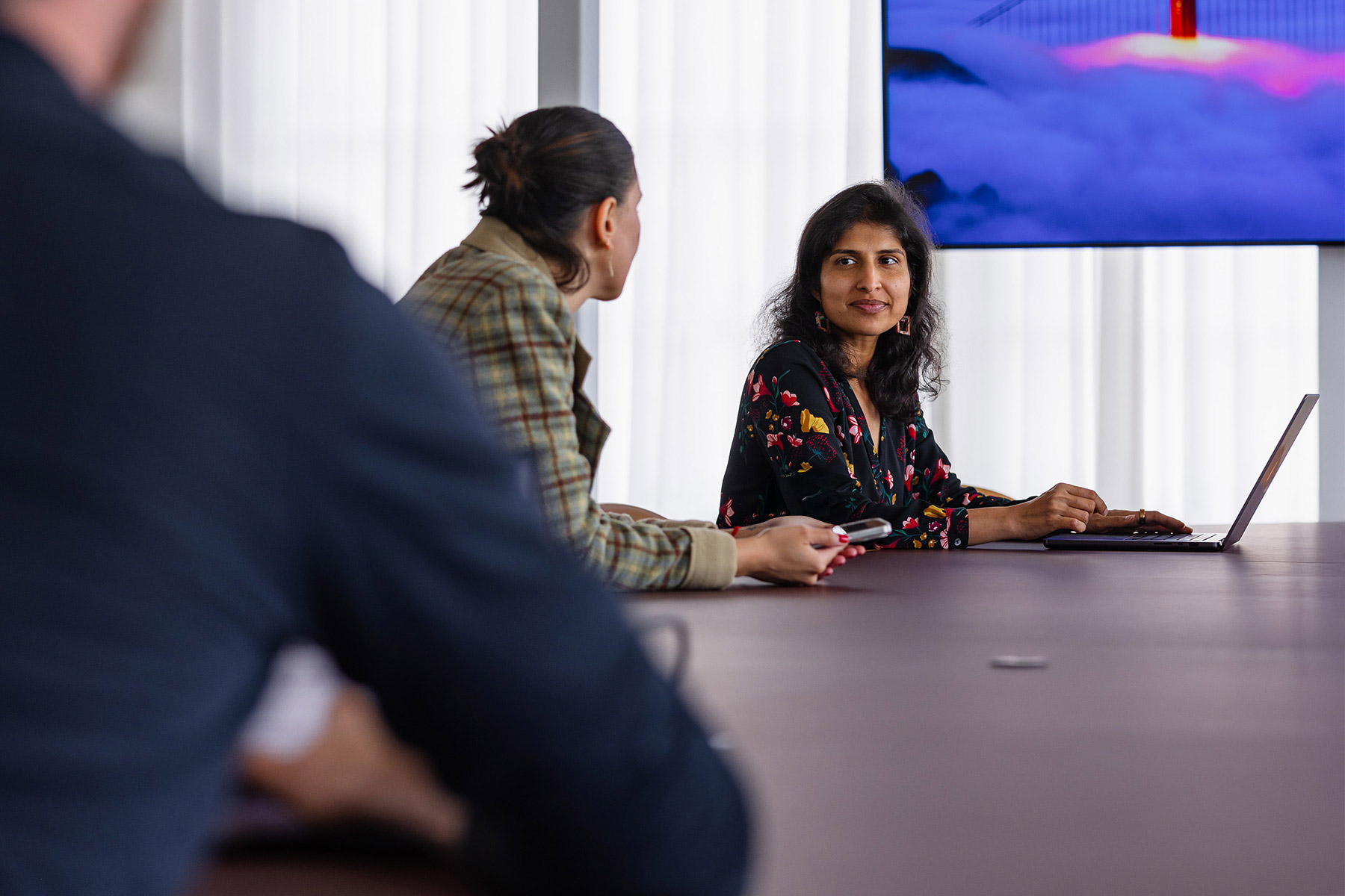 Two women engaged in a discussion at a meeting table, one using a laptop, with a large screen displaying visuals in the background.