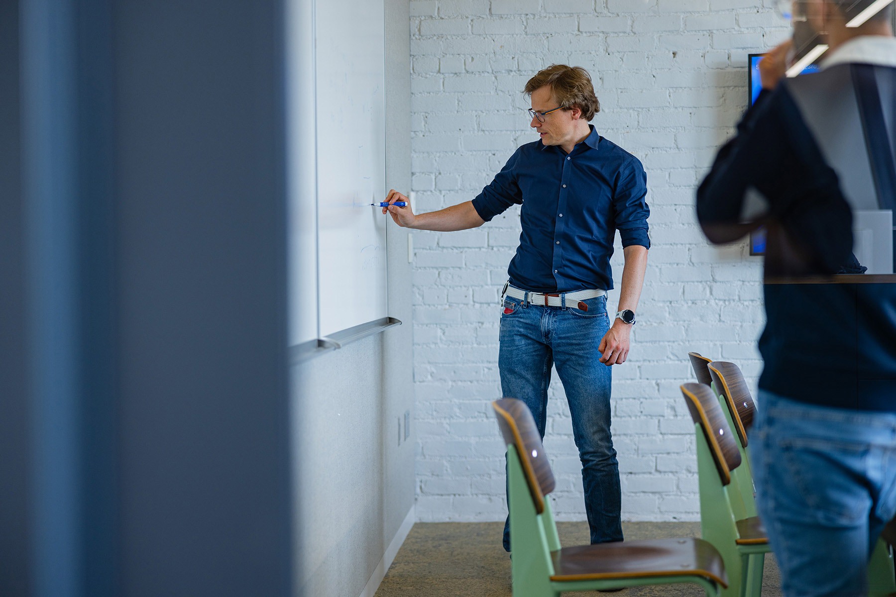 A person standing at a whiteboard, writing notes during a presentation in a bright room with chairs in the foreground.