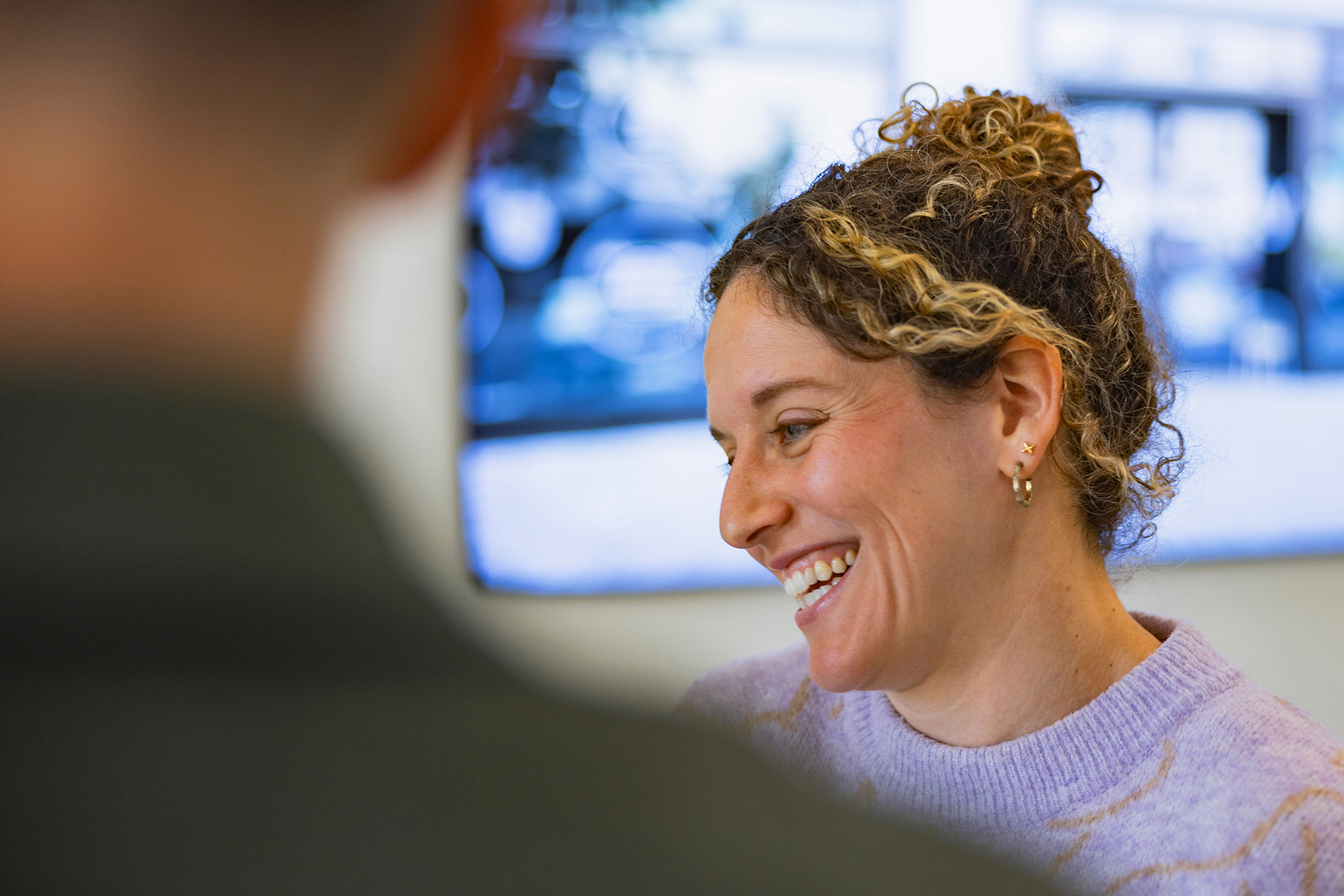 A woman with curly hair smiles while engaged in conversation, demonstrating a positive interaction that emphasizes community and connection.