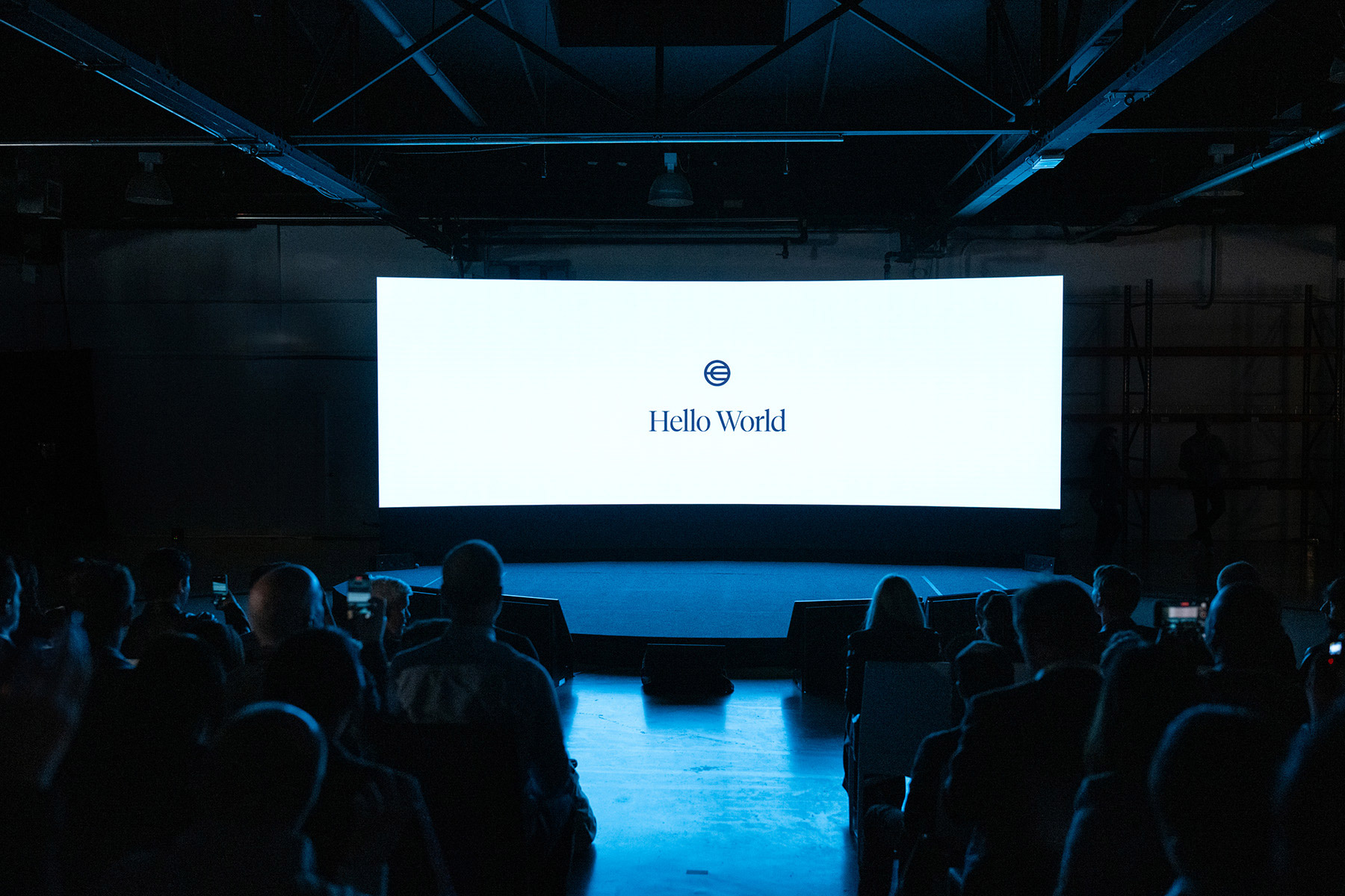 A stage set for a presentation, featuring a large screen displaying 'Hello World' and the World symbol, with an audience in silhouette.