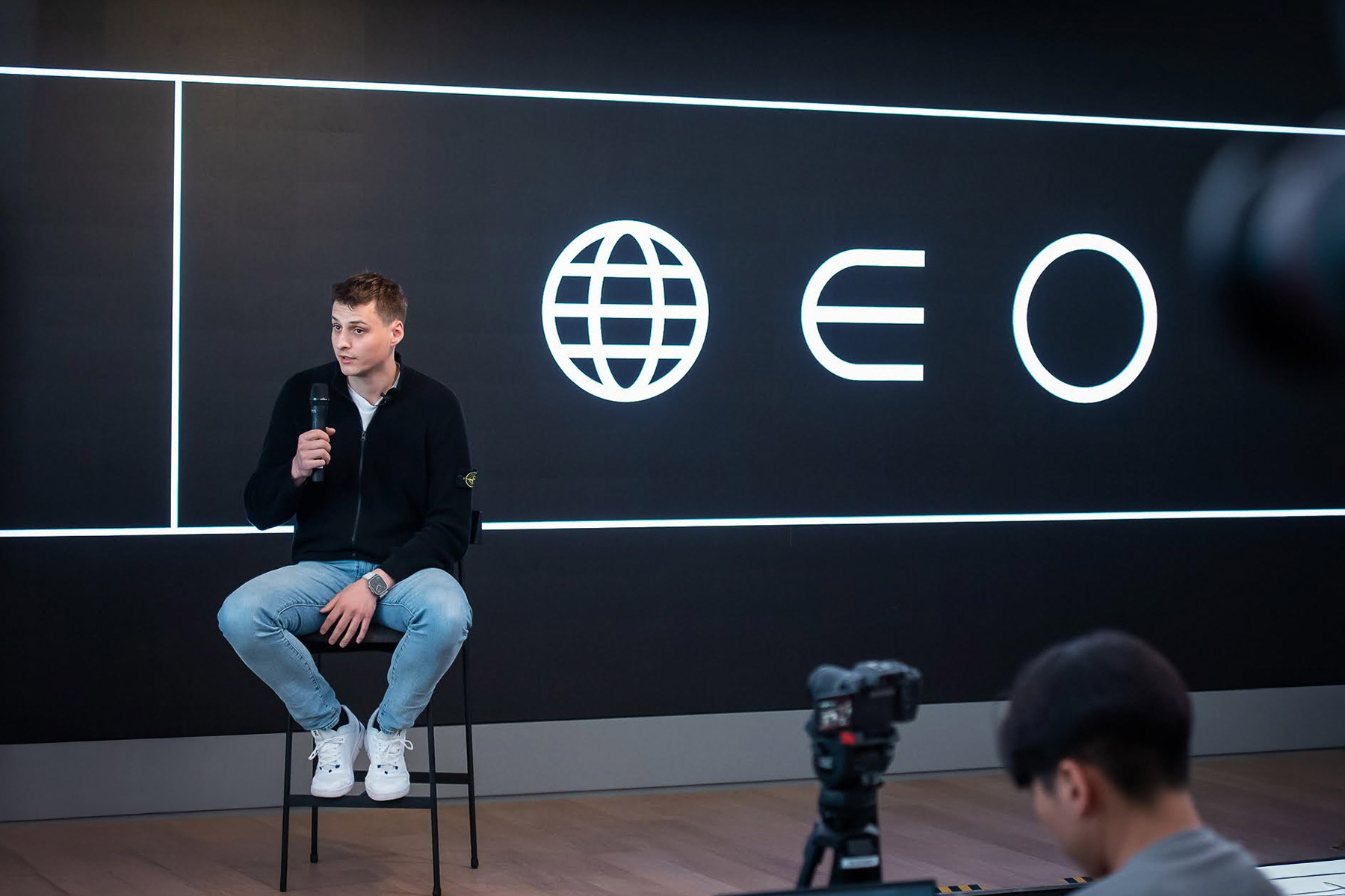 A speaker sitting on a stool in front of a large screen displaying the new Worldcoin logo, emphasizing innovation and community engagement.