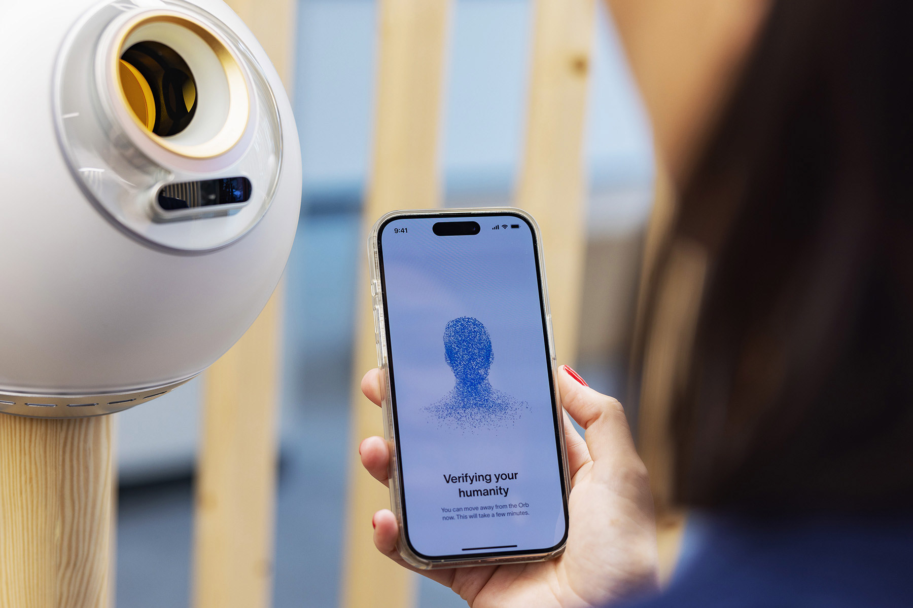 A user verifies their identity using the Orb while holding a phone displaying a biometric verification prompt.