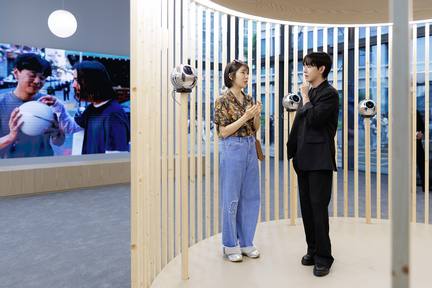 Two individuals engaging in conversation near biometric verification Orbs, with a digital screen displaying a community scene in the background.