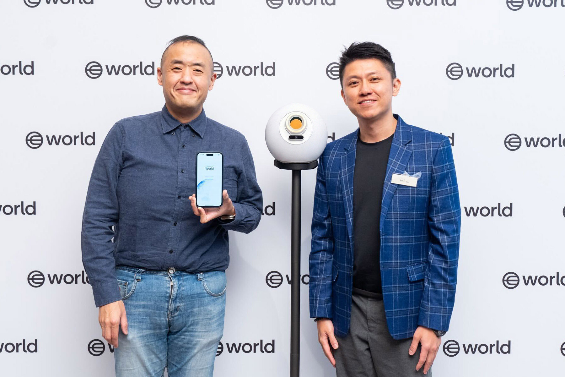 Two individuals stand in front of a World-branded backdrop, one holding a smartphone displaying the World App, next to an Orb on a stand.