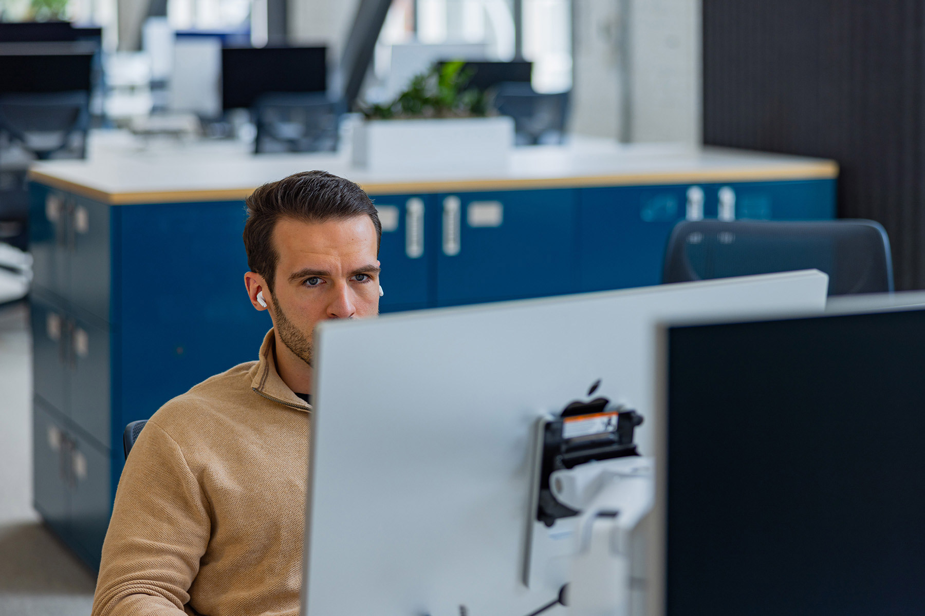 A focused individual wearing earbuds works on a computer in a modern office environment with blue storage cabinets.