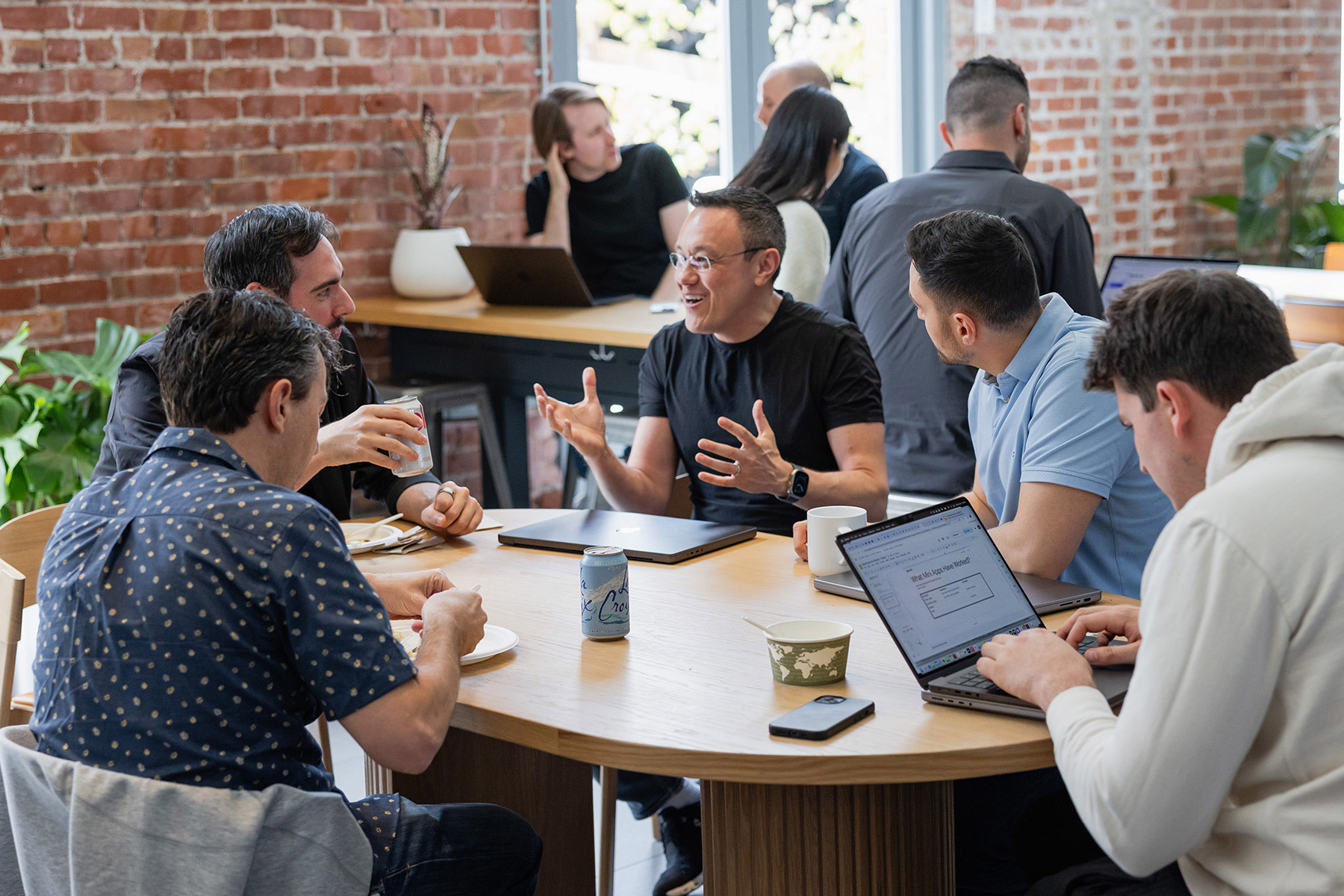 A collaborative meeting with diverse individuals discussing ideas around a table, surrounded by laptops and plants.