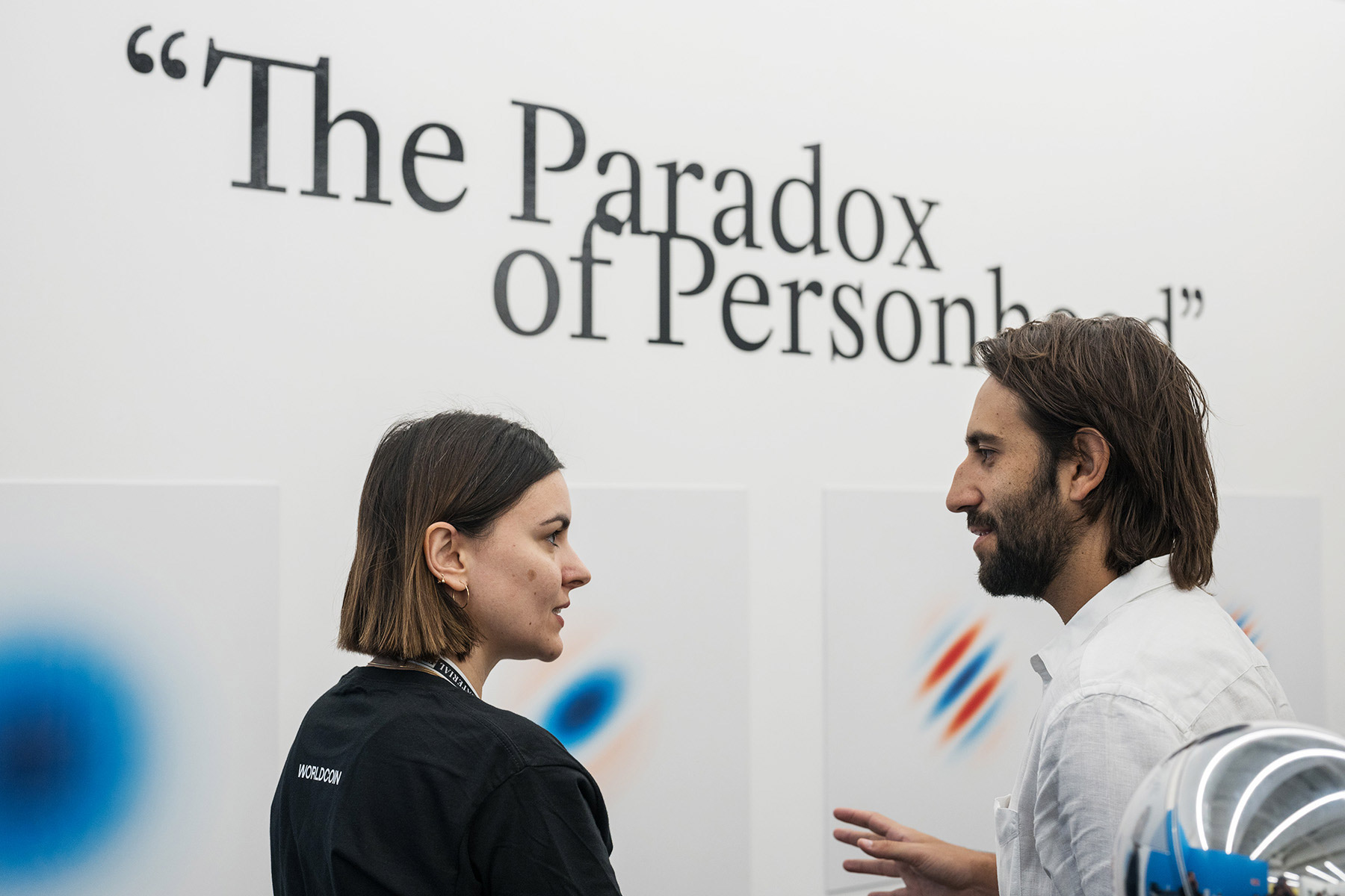 Two people engaging in a conversation in front of a wall displaying the phrase 'The Paradox of Personhood.'
