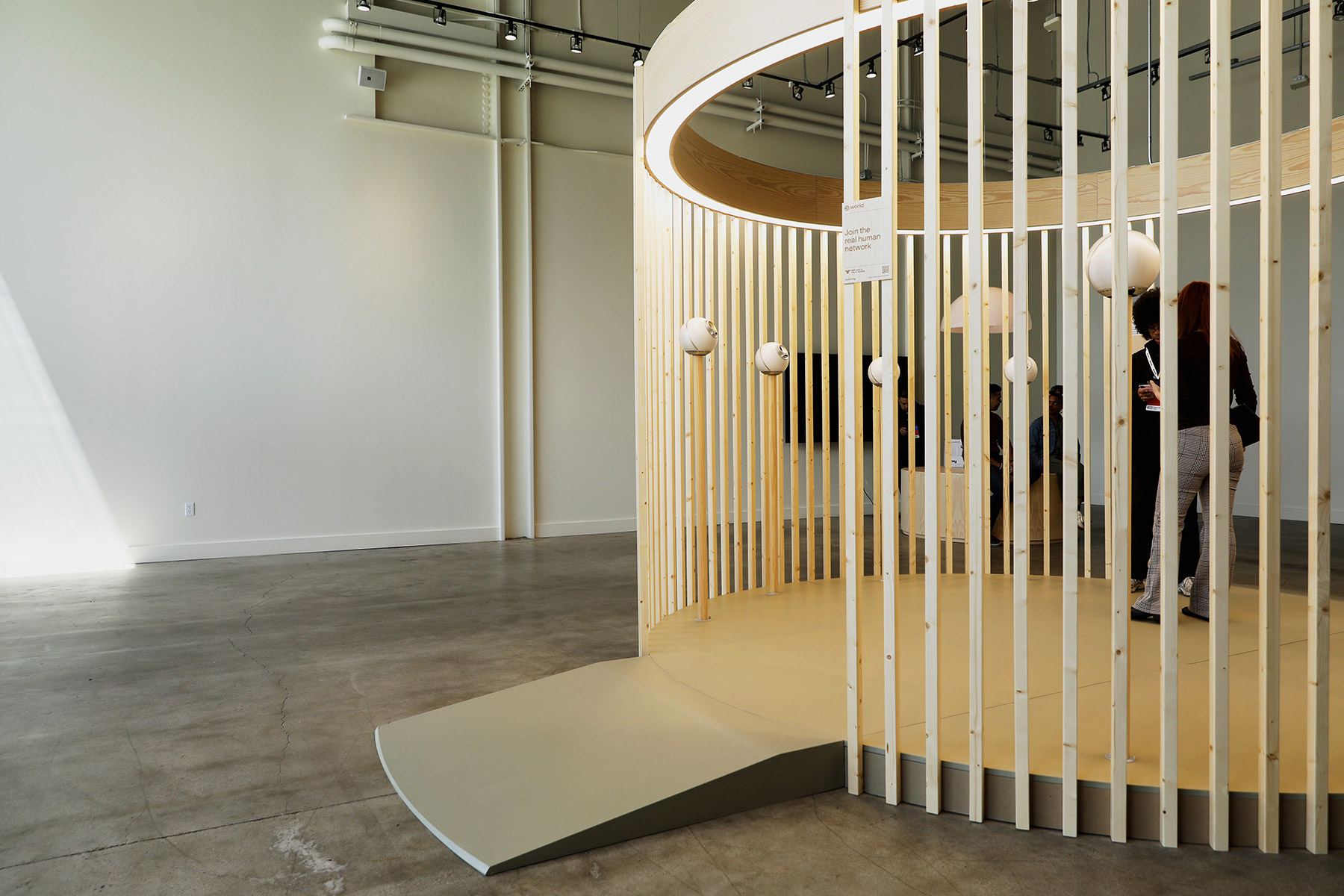 A circular wooden structure with vertical slats, containing spherical Orbs for biometric verification. People are interacting with the setup in a well-lit space.