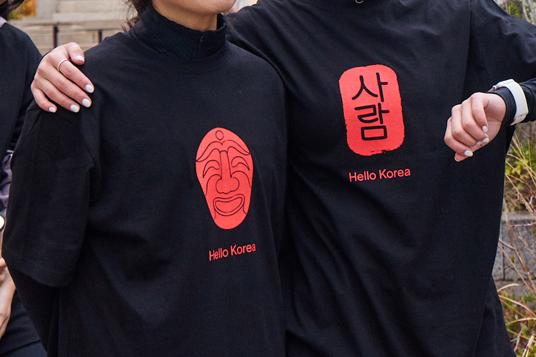 Two individuals wearing black shirts with red designs, one featuring a face mask and the other saying 'Hello Korea' in Korean and English.