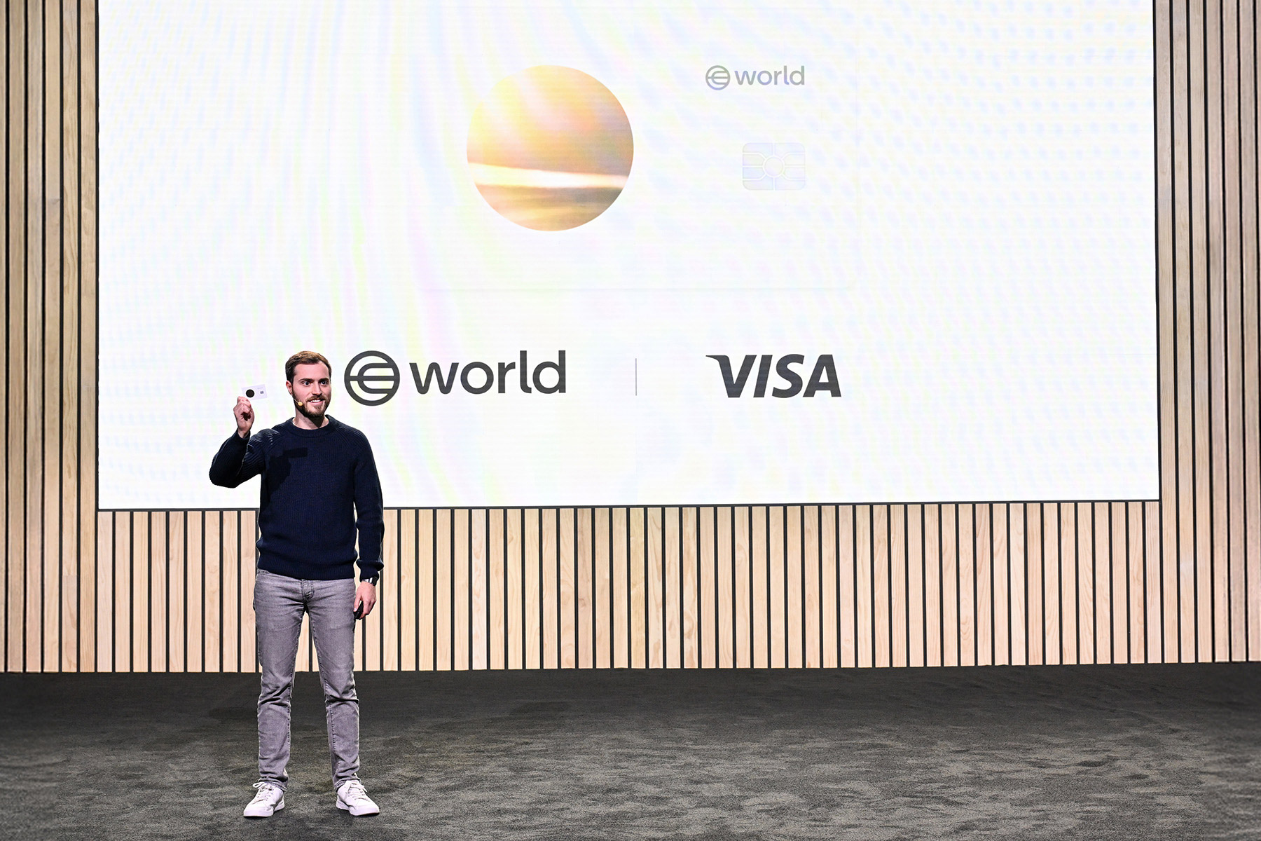 A speaker presenting on stage with a screen behind displaying the World and Visa logos, highlighting digital asset integration.