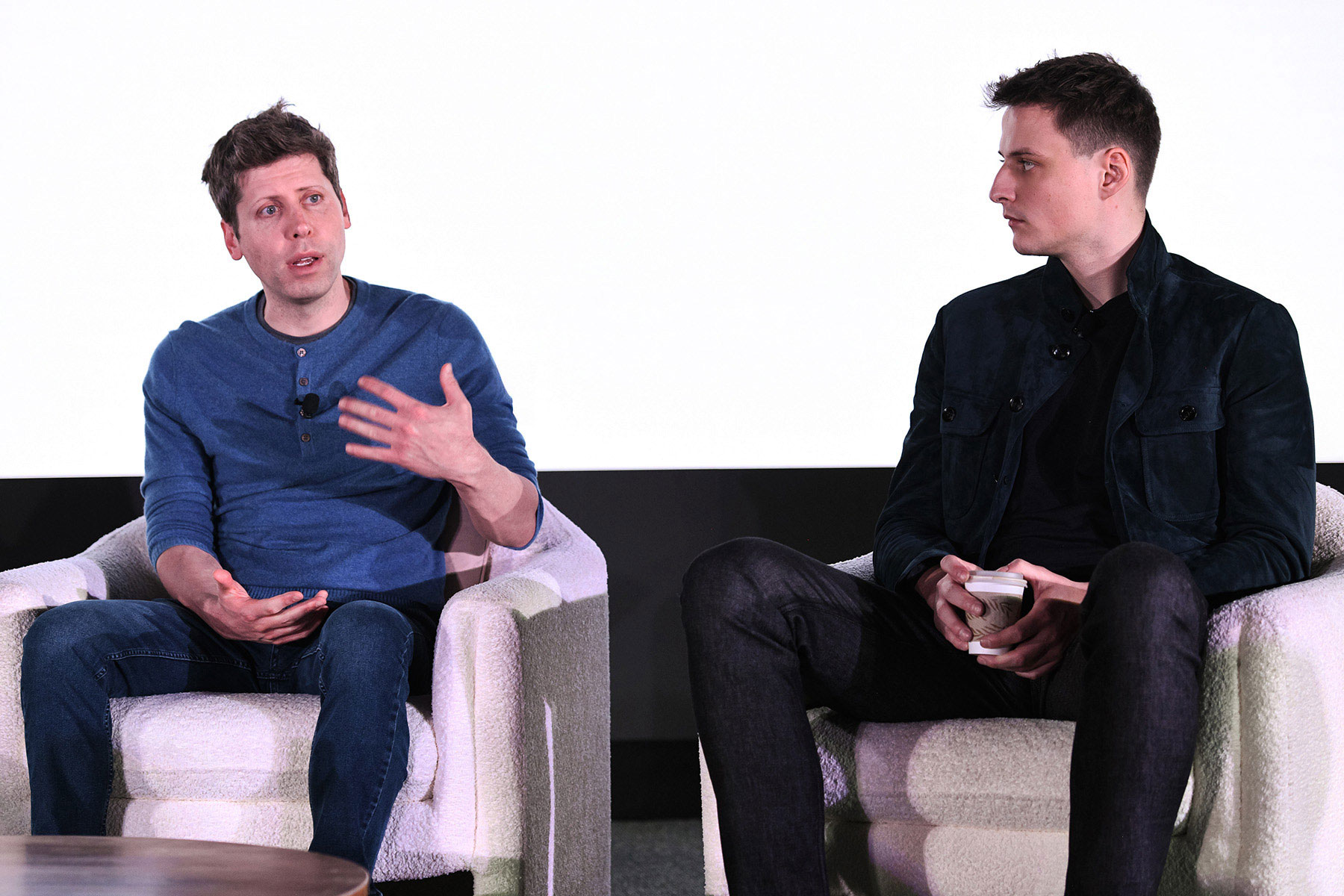 Two speakers engage in conversation on stage, discussing enhancements to World's Bug Bounty Program. One speaker gestures while the other listens attentively.
