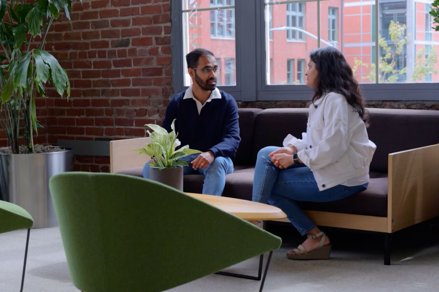 Two individuals engage in a conversation on a couch in a modern, well-lit space, surrounded by greenery.