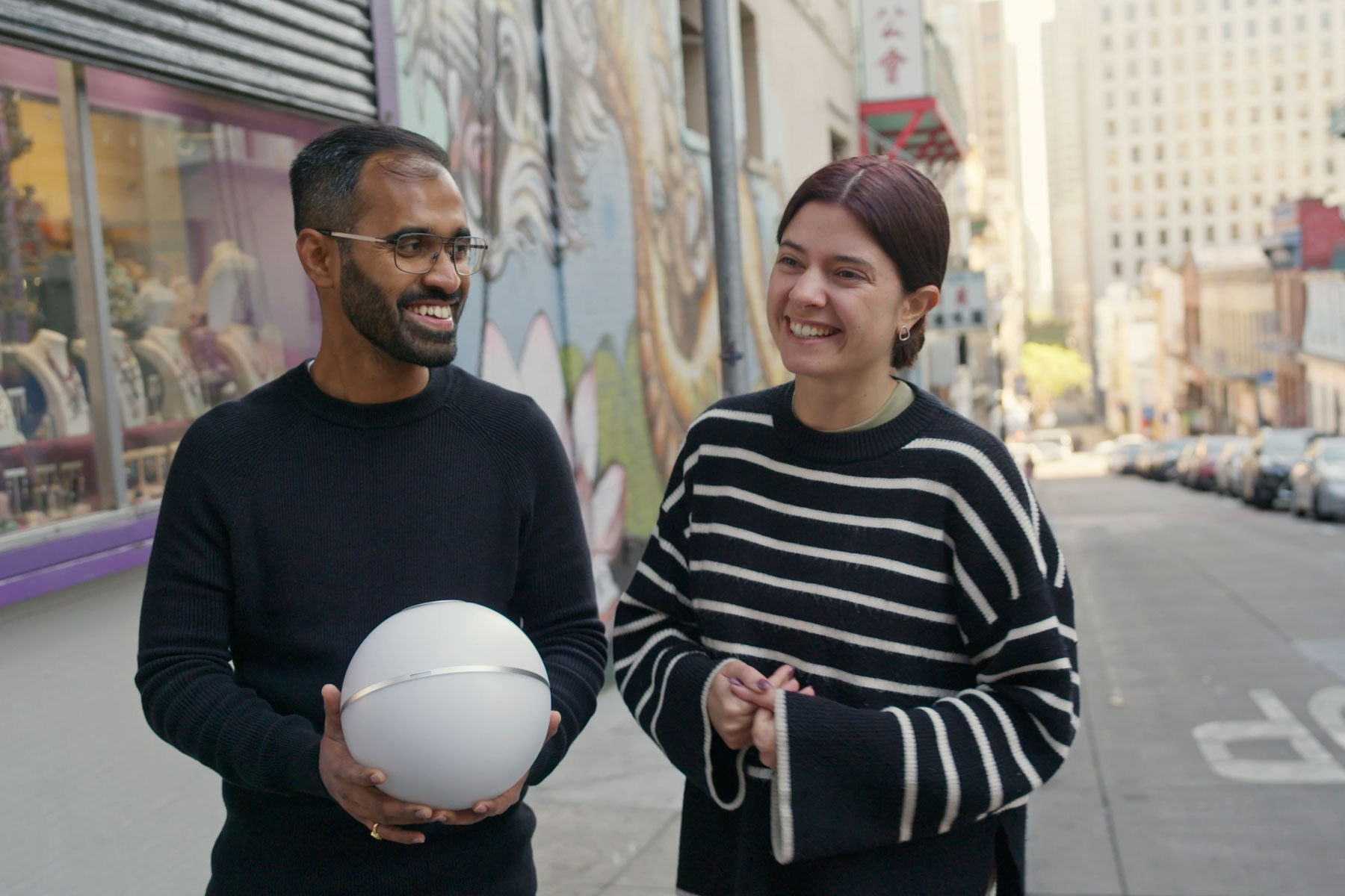 Two people are smiling and engaging while holding the Orb on a city street, with colorful murals in the background.