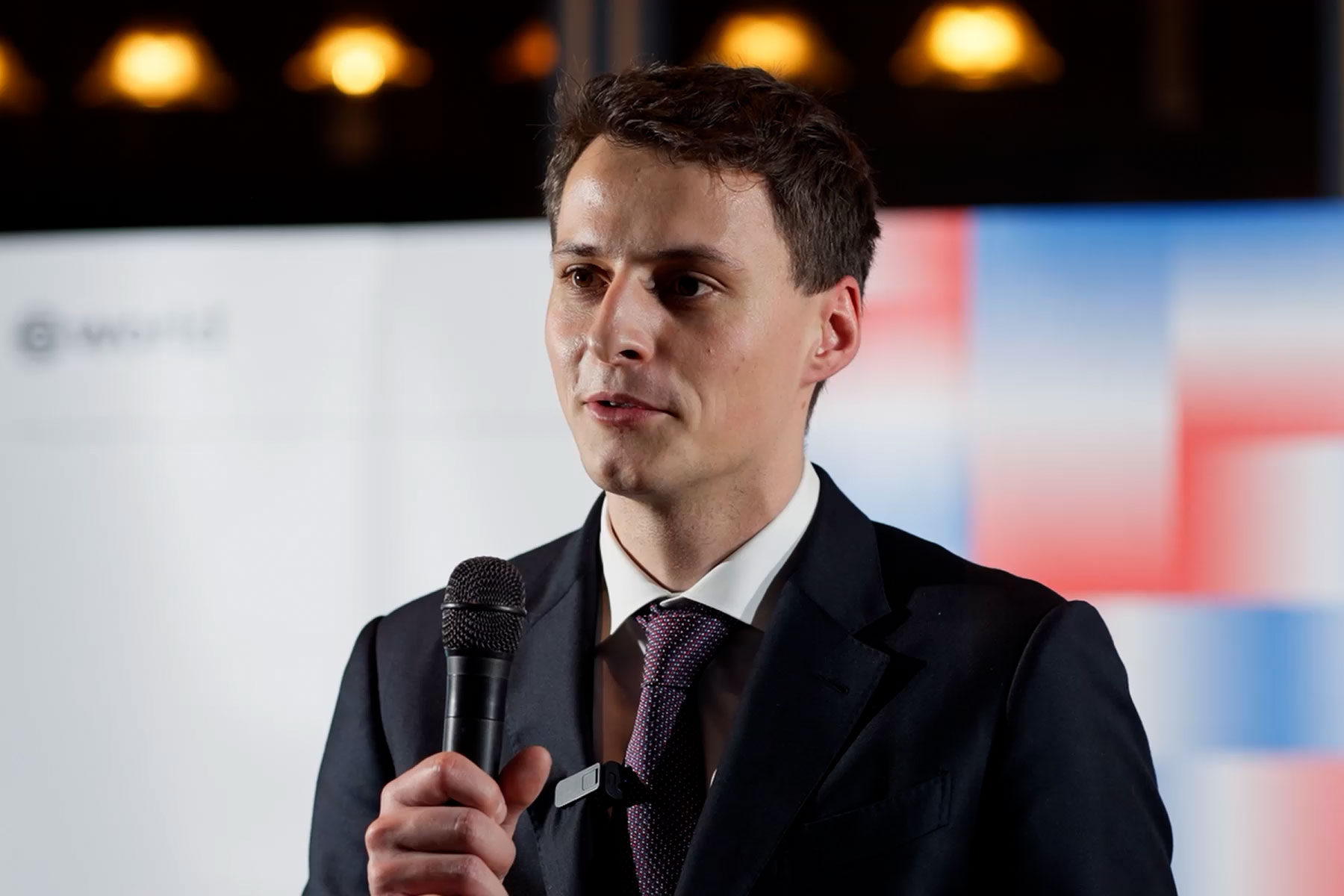A man in a suit speaks into a microphone at an event, with a World logo in the background and colorful visual elements.