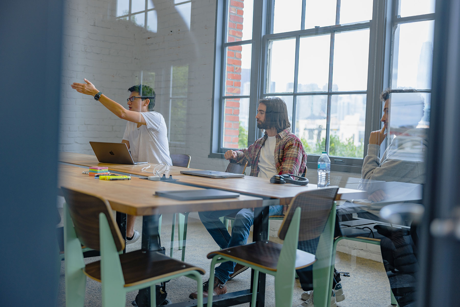 Three people in a meeting room discussing ideas around a table with a laptop and notes. Large windows show a cityscape outside.