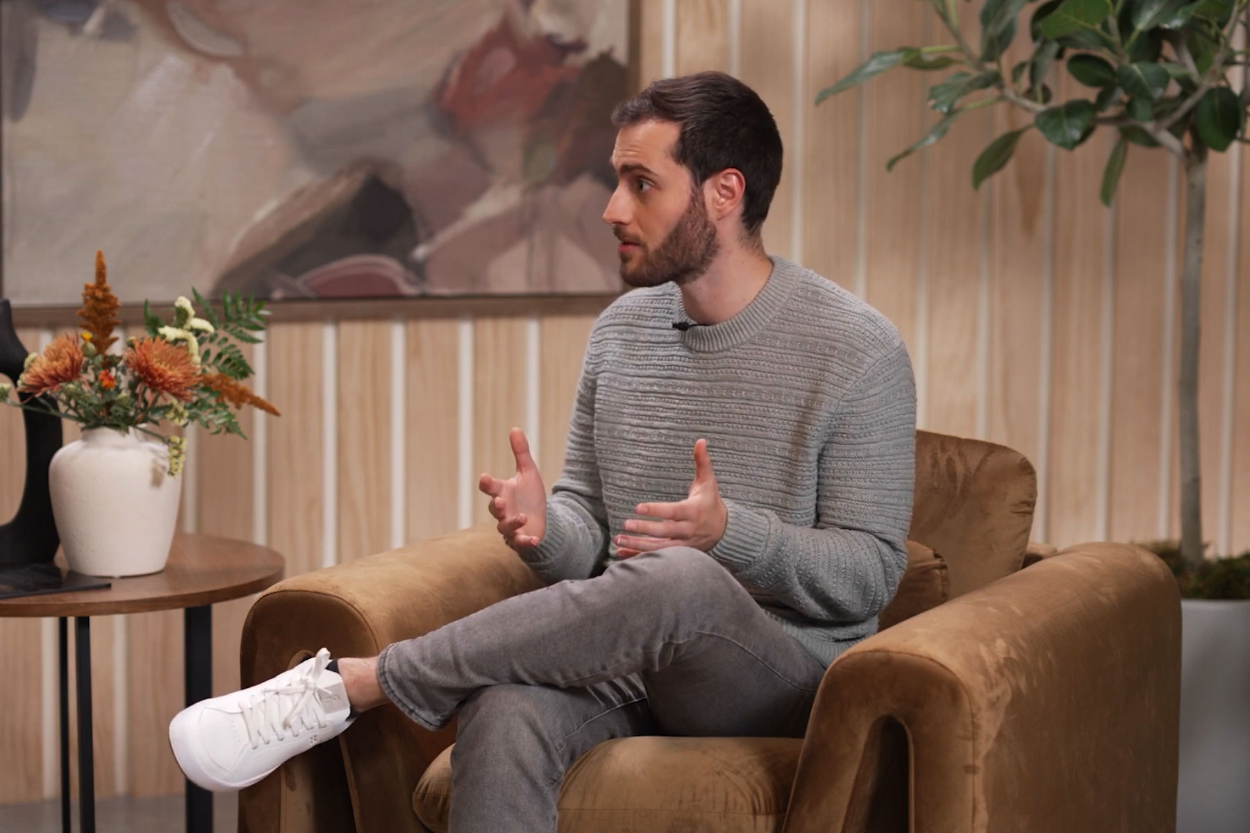 A person sitting on a brown chair, speaking animatedly, with a plant and artistic backdrop, discussing Worldcoin.