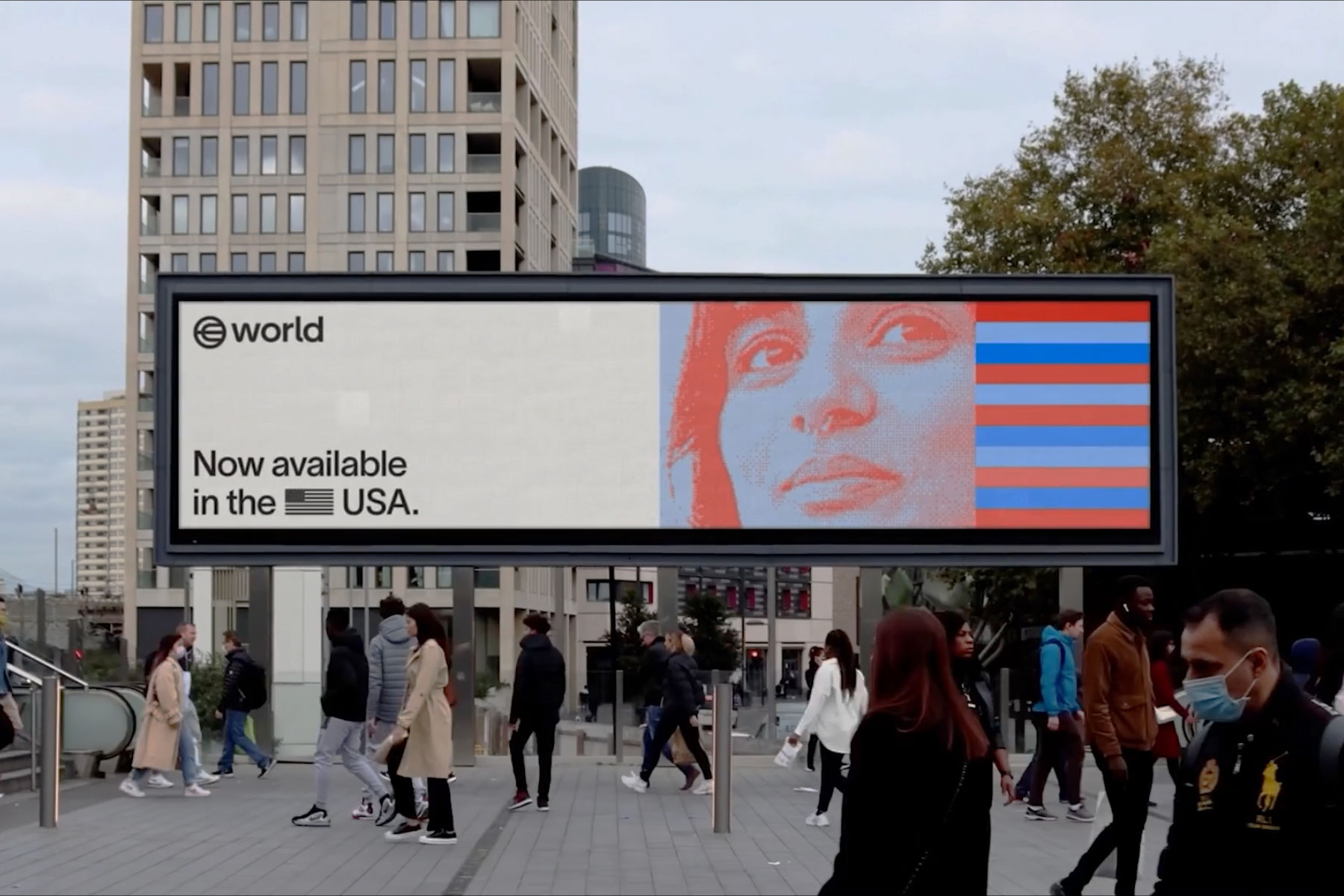 A digital billboard displays a promotional announcement for World, featuring a colorful illustration of a person's face and text reading 'Now available in the USA.' People walk by in an urban setting.