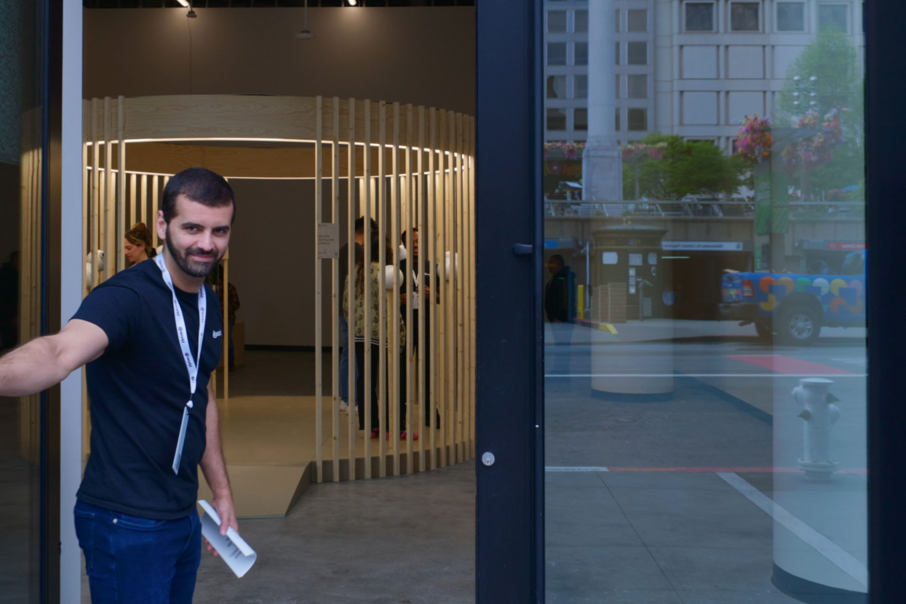 A person stands at the entrance of a modern World storefront, welcoming visitors with a smile. Inside, a wooden structure resembling the Orb is visible, hinting at the technological experience inside.