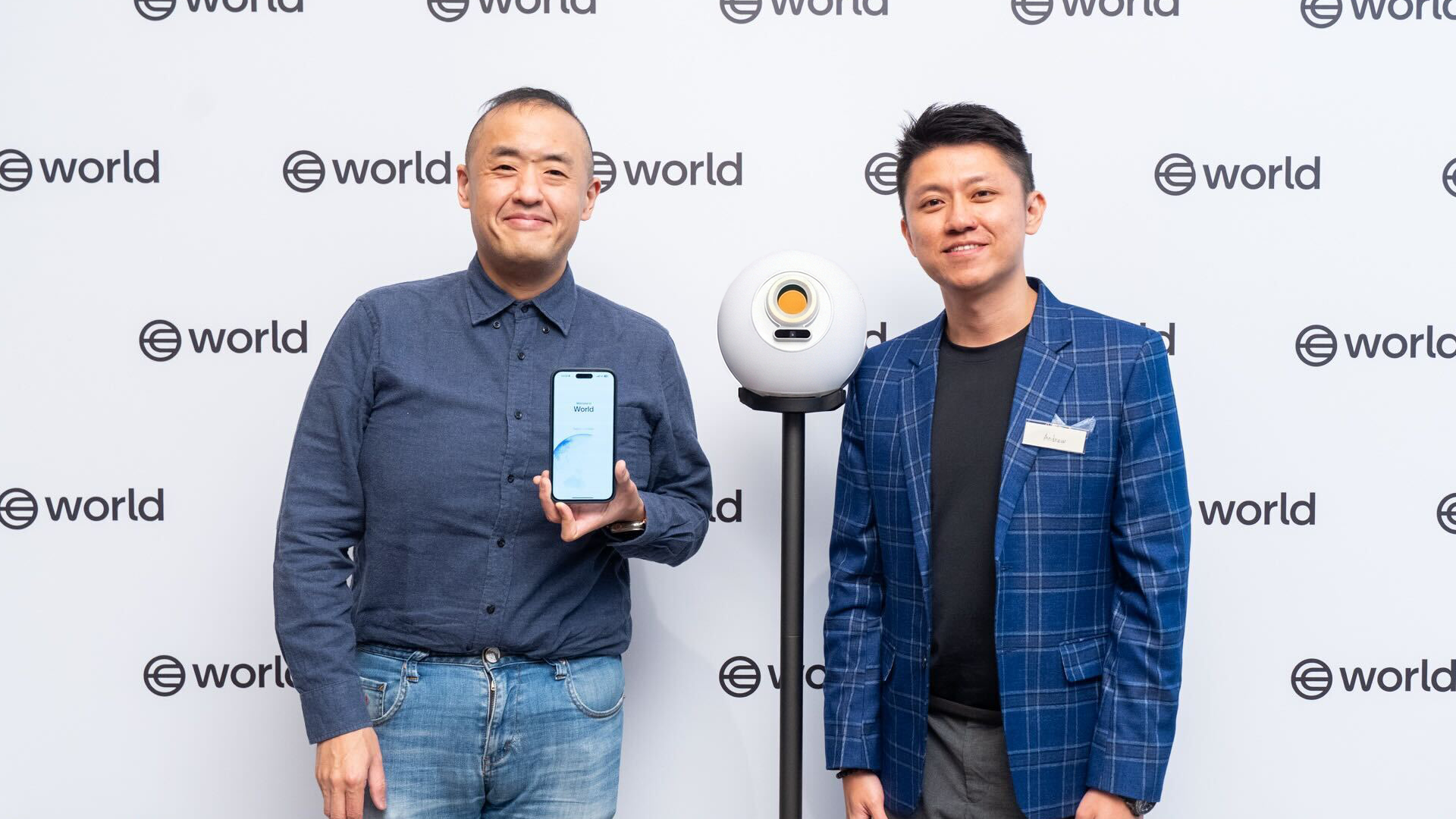 Dwie osoby stoją obok Orb i smartfona, prezentując technologię biometrycznej weryfikacji na wydarzeniu World.