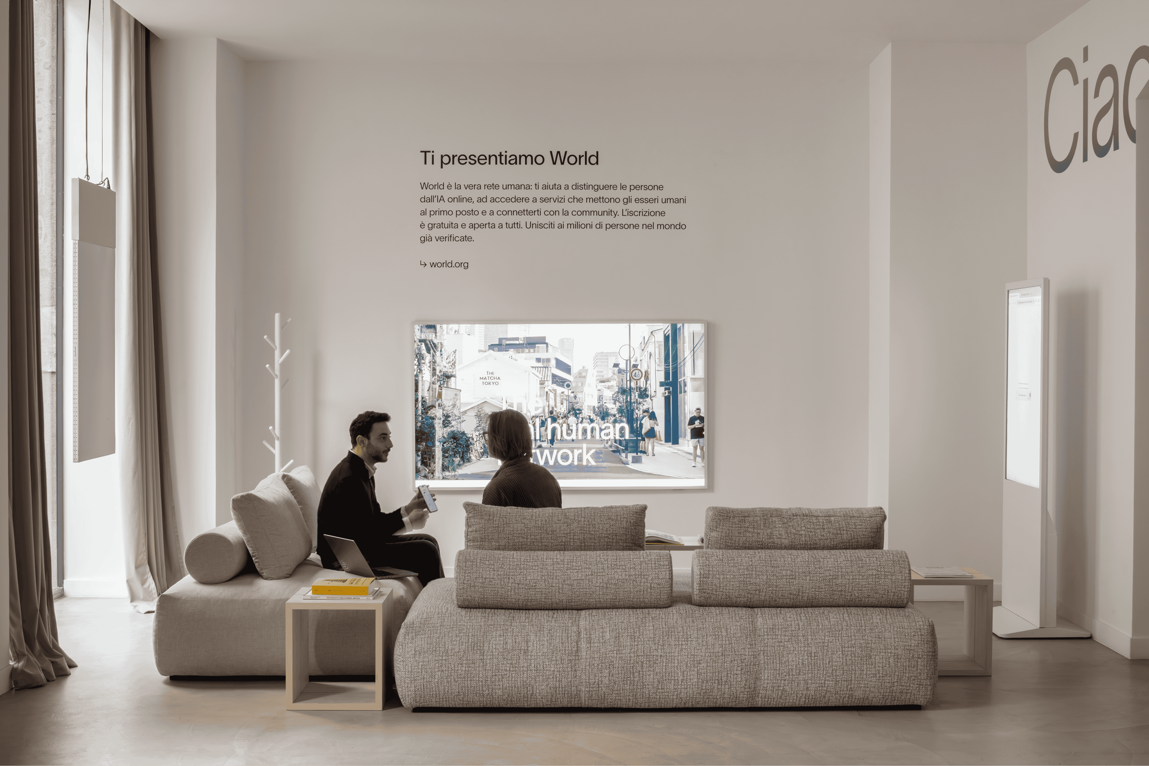 Dos personas conversando en una sala moderna y elegante con una gran pantalla que muestra el texto 'Ti presentiamo World' y un ambiente acogedor.