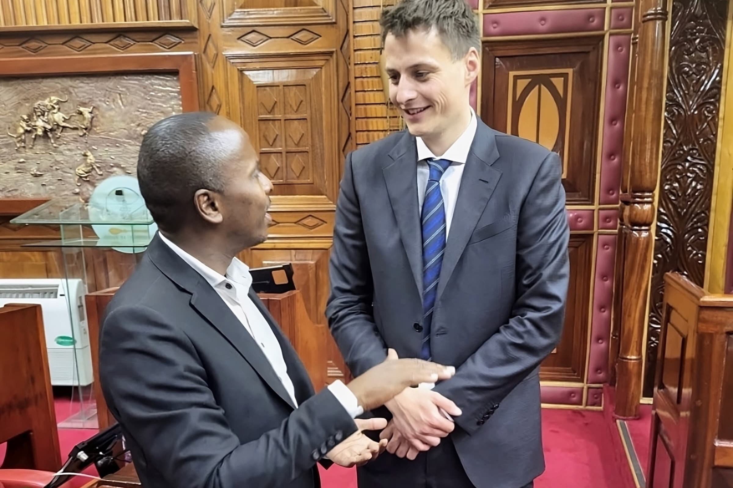 Two men engaging in conversation in a parliamentary setting, with wooden decorative elements in the background.