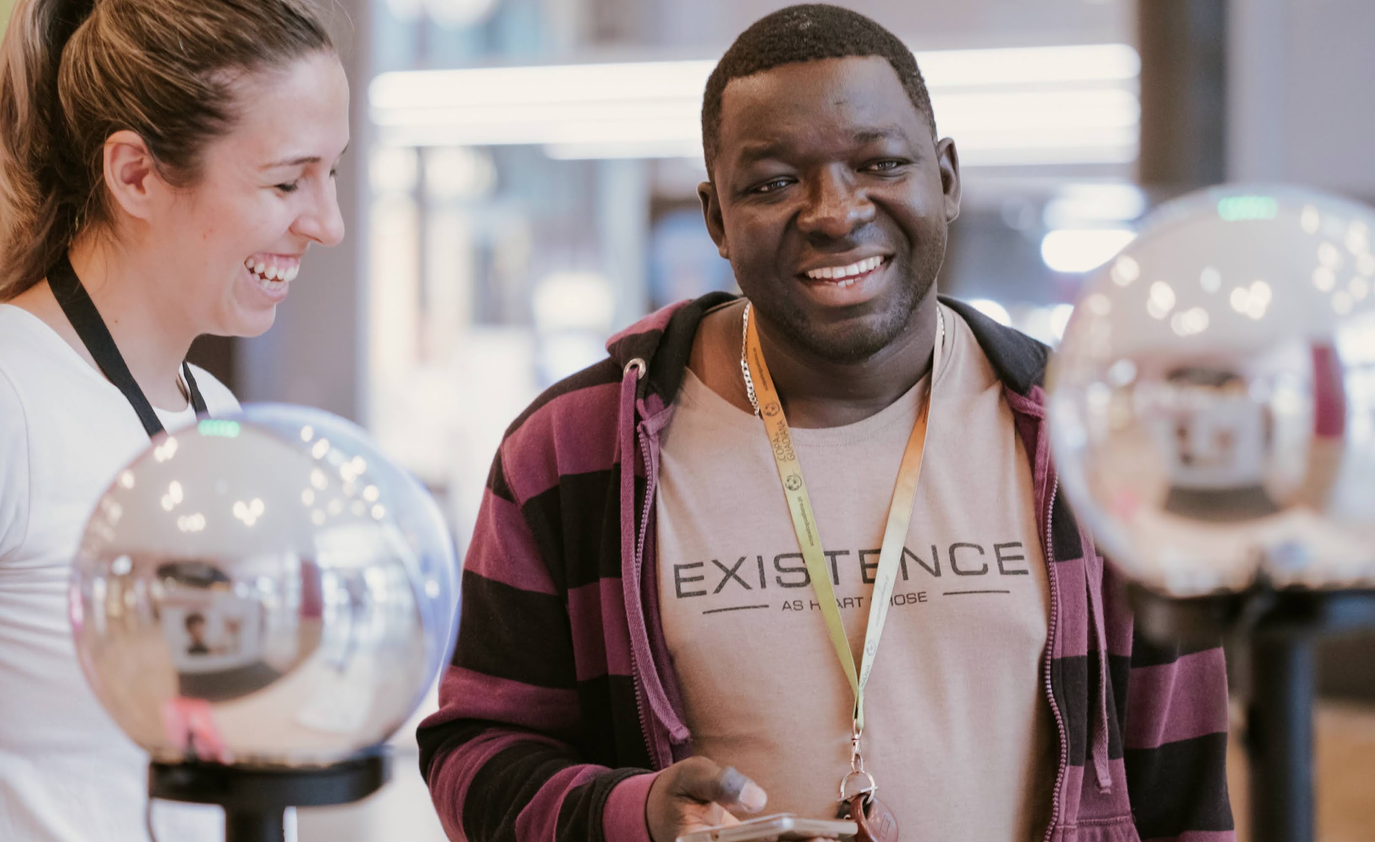 A man smiles while using a smartphone near two Orbs, as a woman laughs in the background, engaged in conversation.
